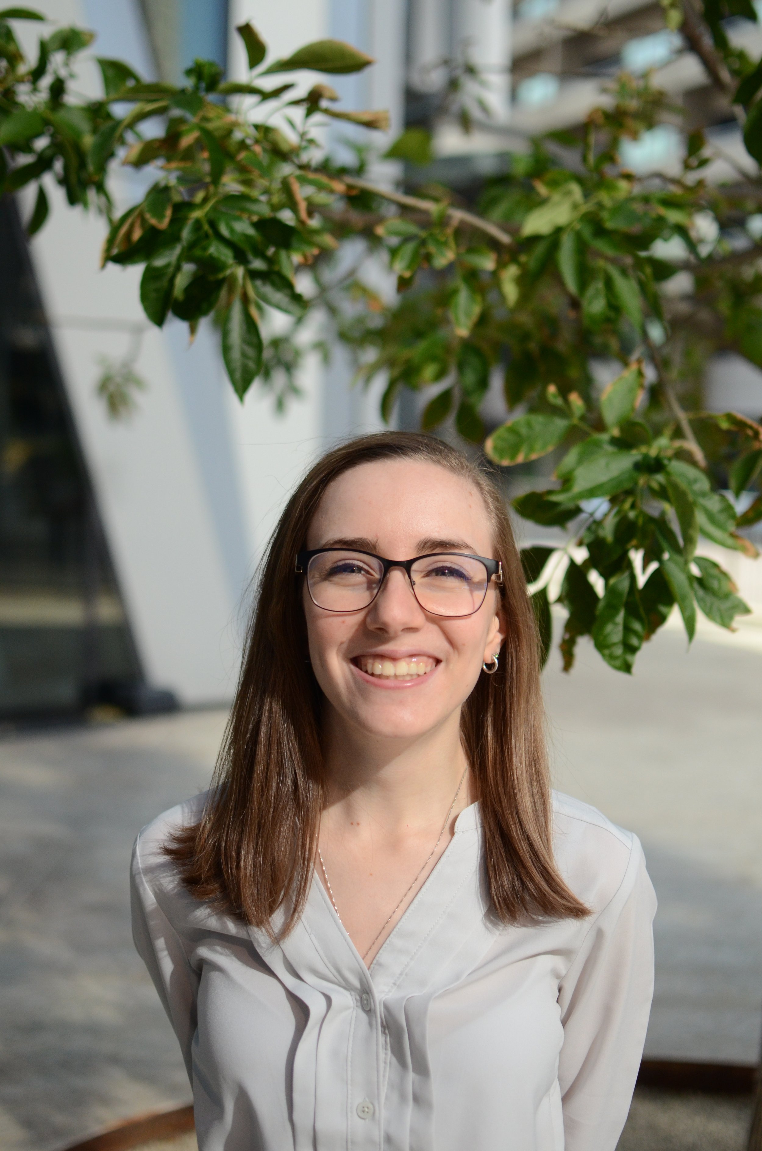 A young woman with glasses smiling outdoors in front of a green leafy tree, wearing a white blouse.