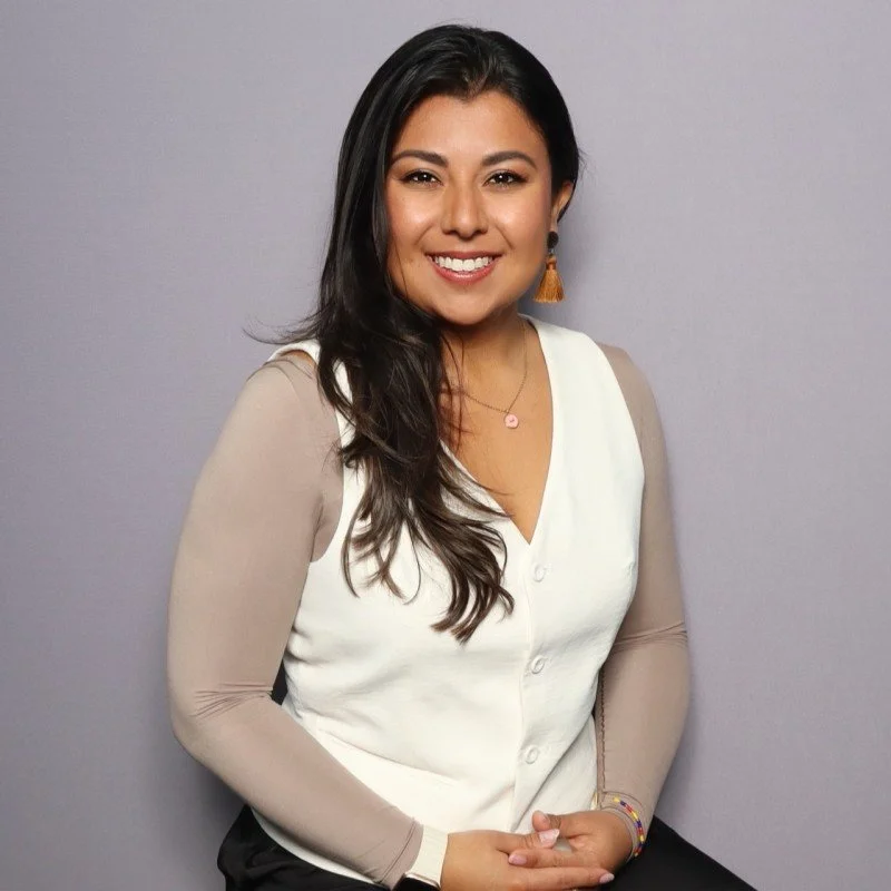 A woman with long dark hair, smiling, wearing a white vest over a beige long-sleeve blouse, with earrings, a necklace, and a colorful bracelet, standing against a plain grey wall.