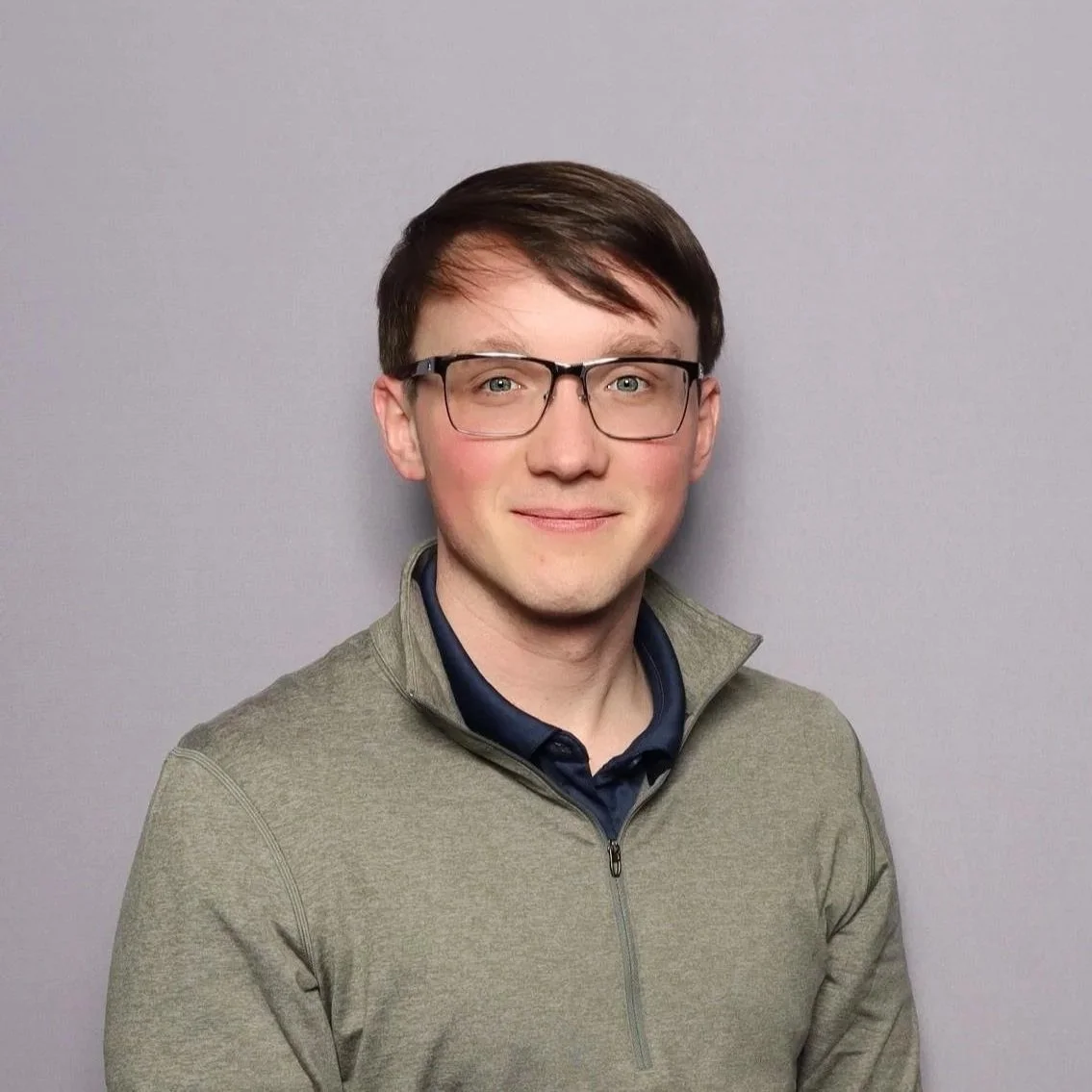 Portrait of a young man with short dark hair, glasses, wearing a gray sweater over a collared shirt, standing against a plain gray background.