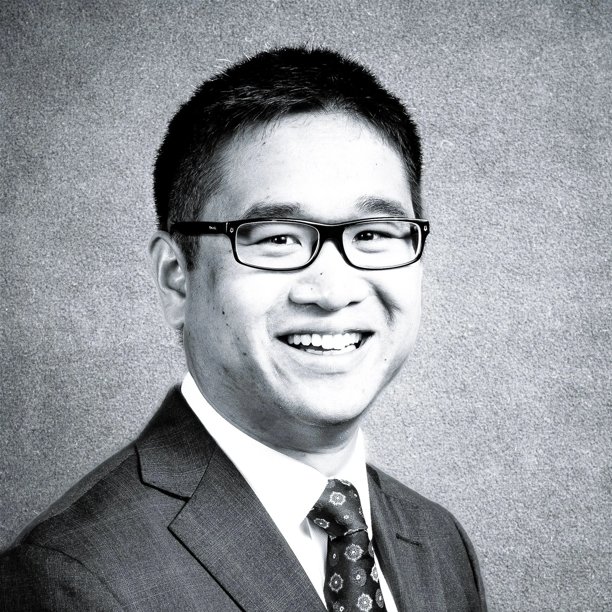 Black and white professional headshot of a smiling man wearing glasses, a suit, a collared shirt, and a patterned tie.