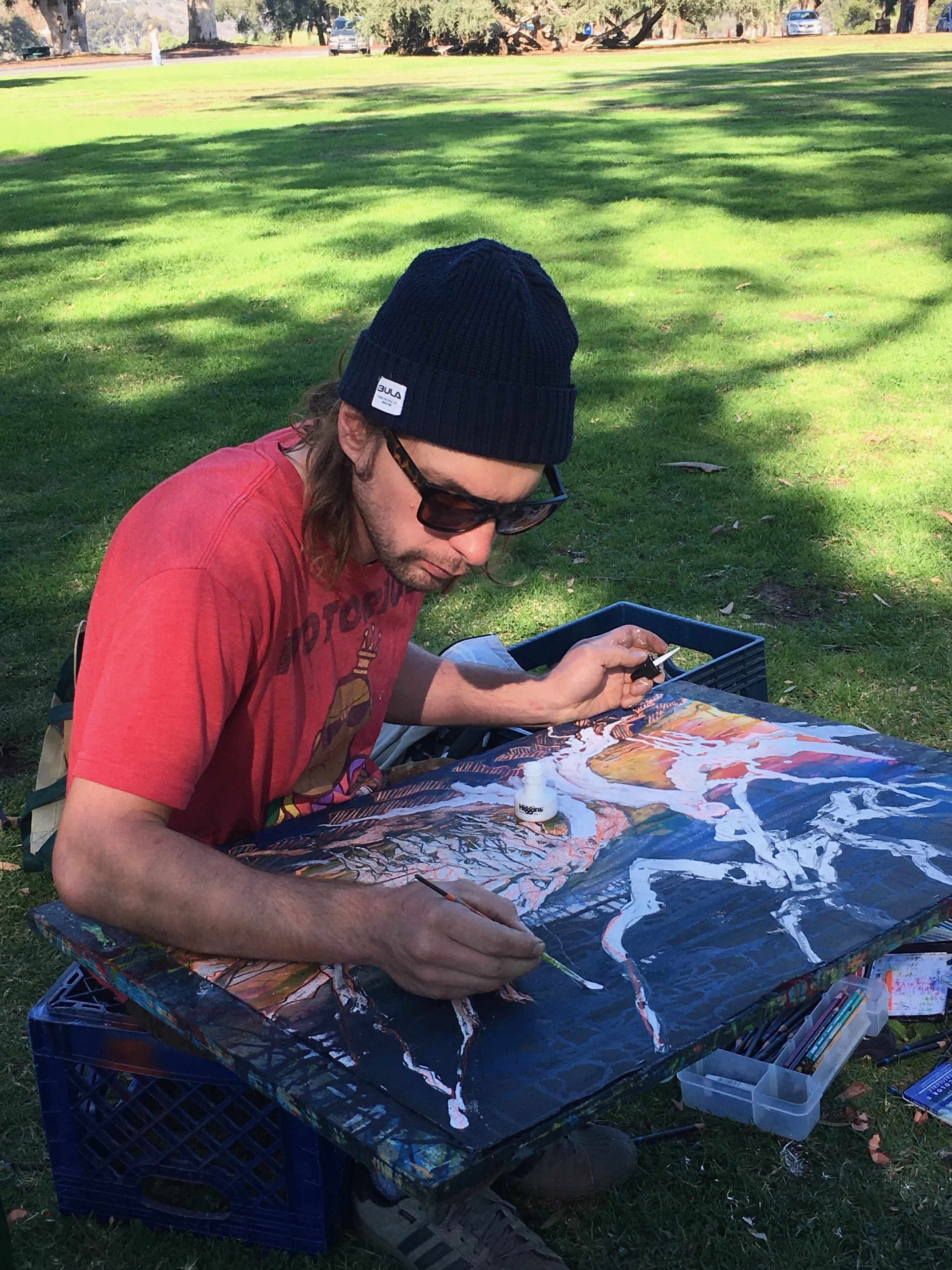 A man wearing a dark blue knit beanie, sunglasses, and a red t-shirt is painting outdoors on a table. The table has a landscape artwork with trees and a dark sky, and art supplies such as paint, brushes, and a small container of white paint. The setting is a park with green grass, trees, and parked cars in the background.