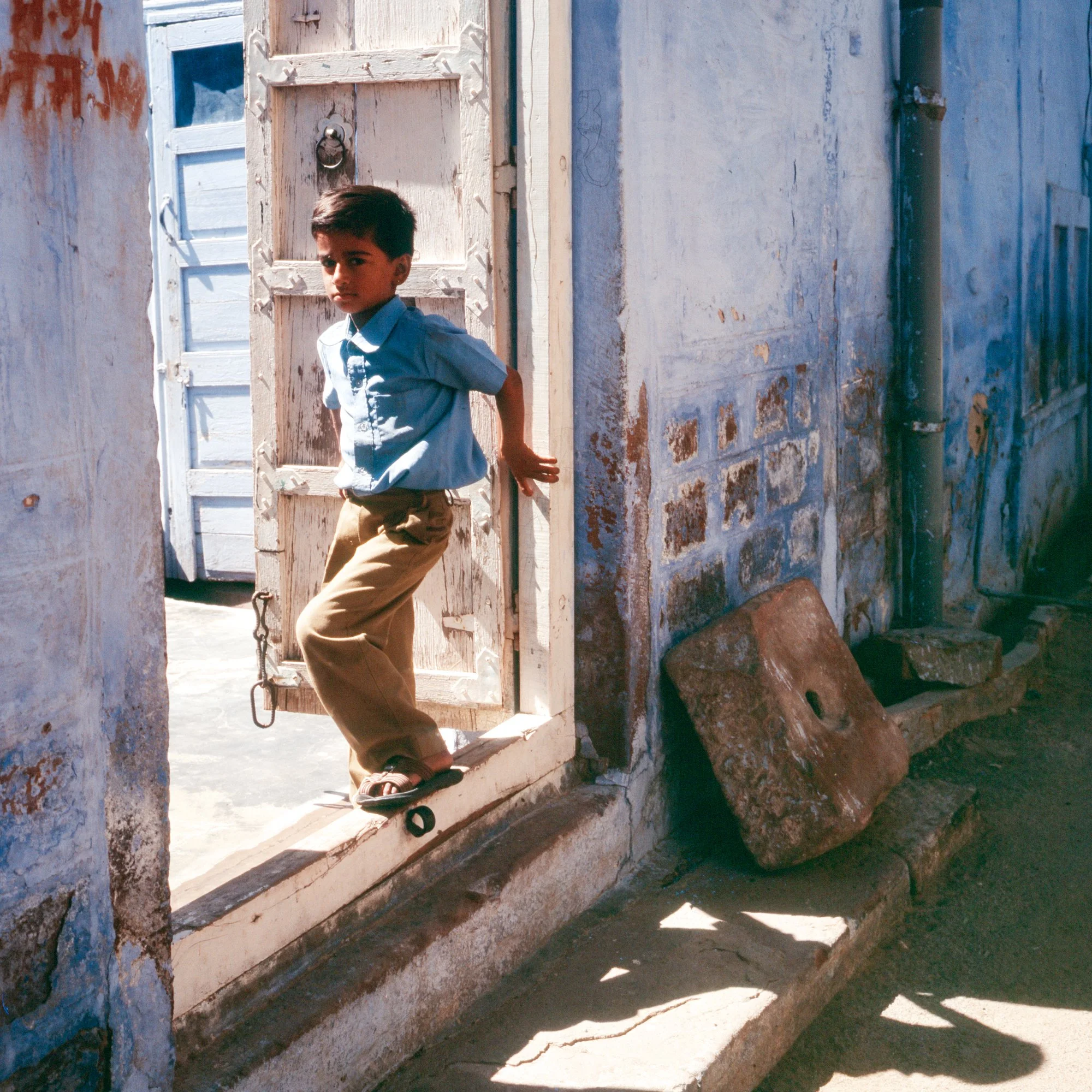 A young boy wearing a blue shirt and tan pants standing on a small ledge at the doorway of a weathered, white-washed building with peeling paint and a rustic wooden door.
