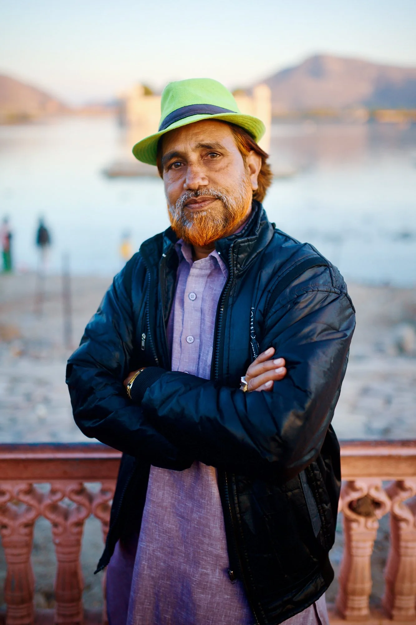 A man with a beard and mustache wearing a lime green hat, a black jacket, and a light purple shirt stands outdoors near a body of water with mountains in the background.