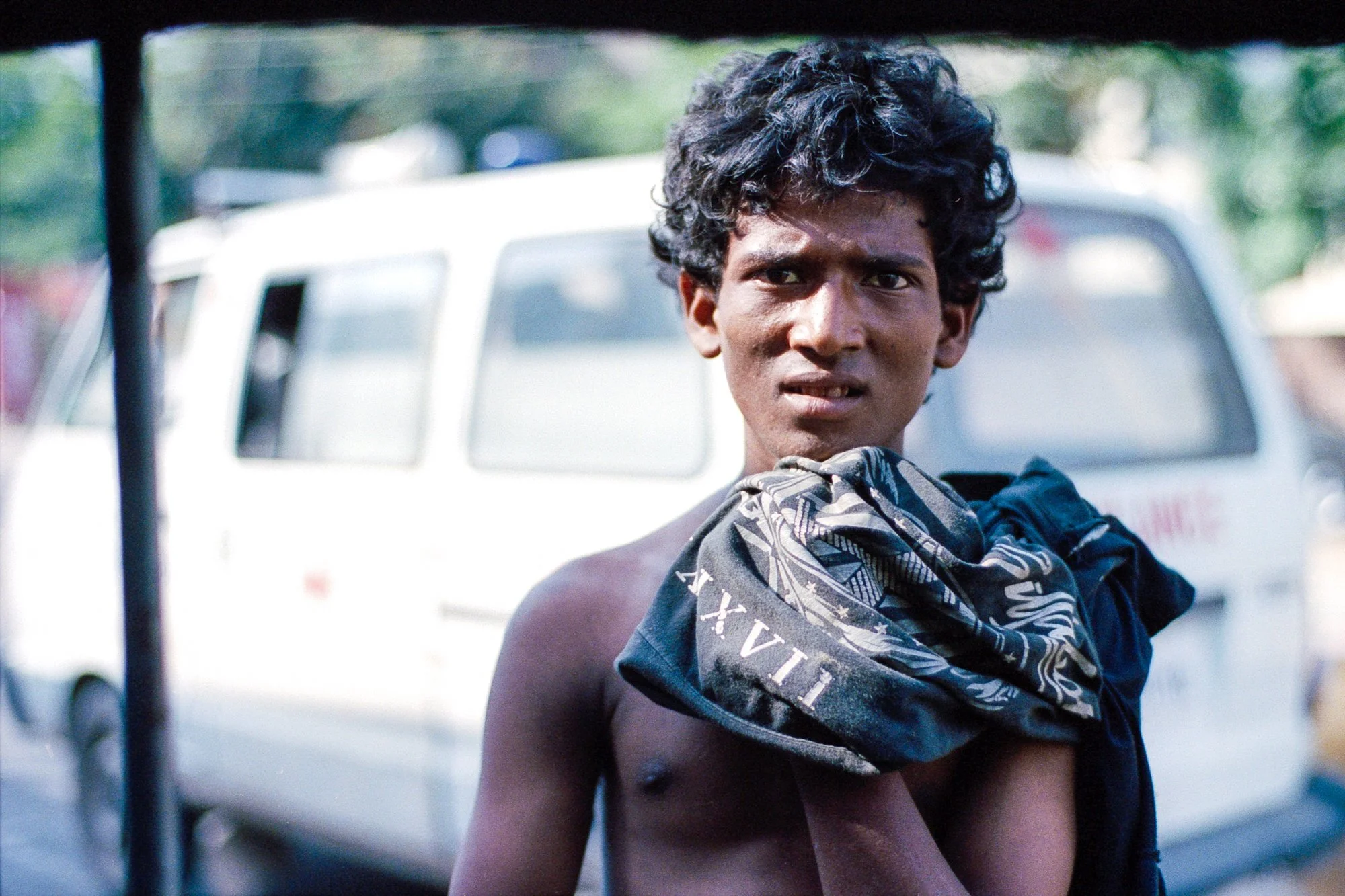 A young man with curly dark hair and a serious expression, holding a cloth or bag over his shoulder, standing outdoors in front of a white vehicle.