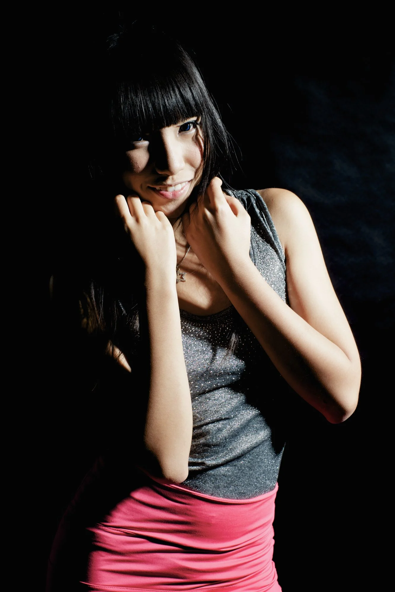 Portrait of a young woman with black hair, smiling, wearing a sparkly gray top and pink skirt, with a dark background.