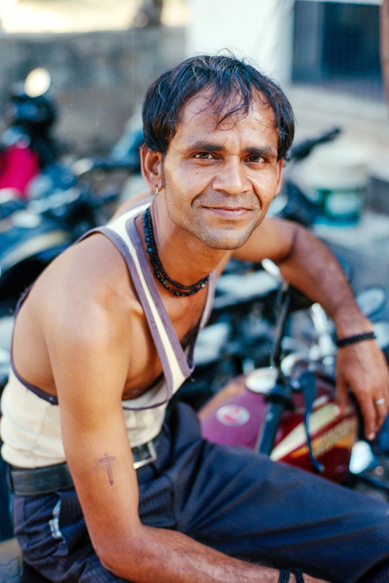A man with short dark hair and a small beard smiling, sitting on a motorcycle outdoors, wearing a tank top and necklaces.