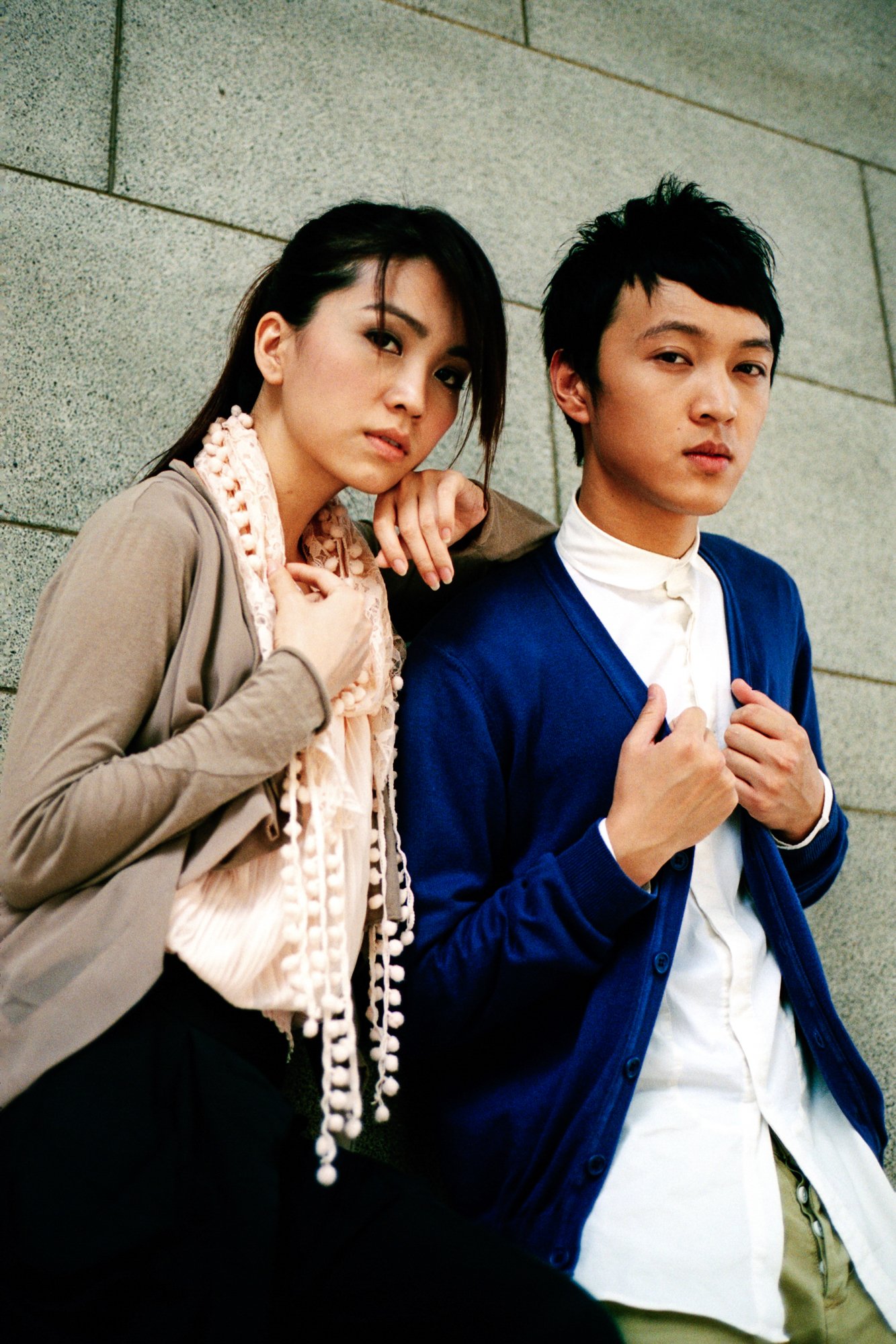 A woman and a man leaning against a gray stone wall, both looking at the camera with serious expressions.