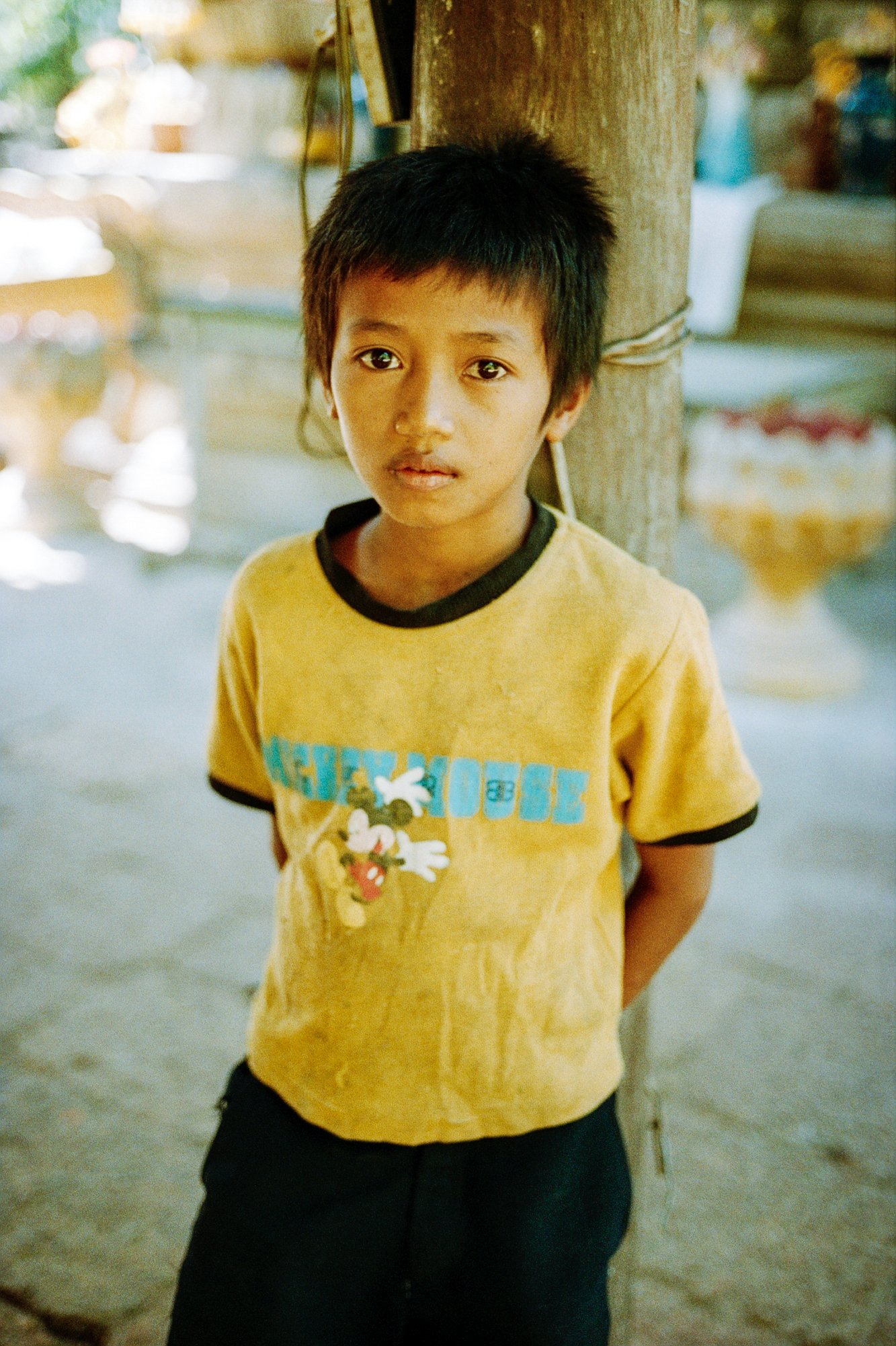 Young boy with short black hair and brown skin standing indoors, leaning against a wooden post, wearing a faded yellow T-shirt with Mickey Mouse on it and black shorts.