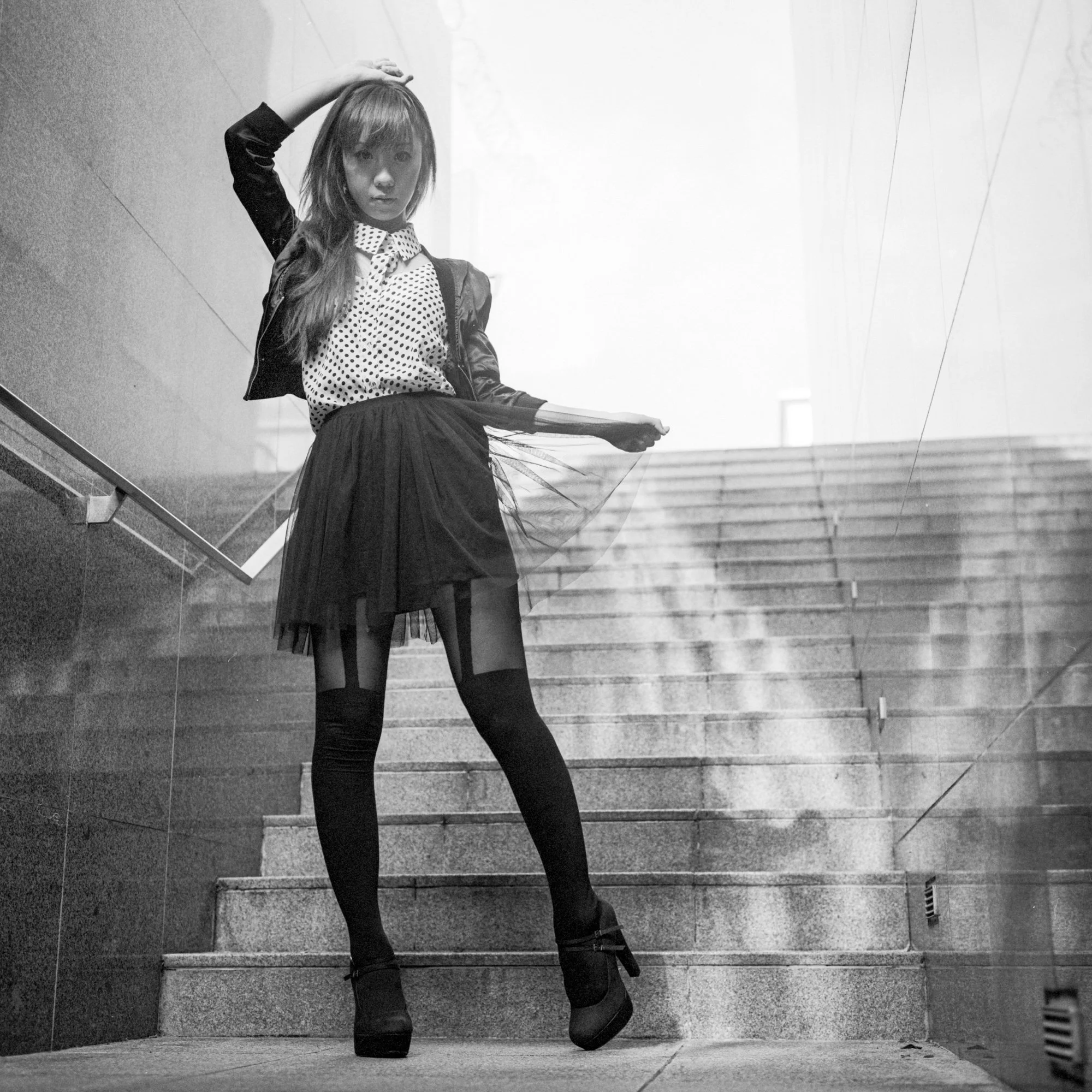 A woman standing on an outdoor staircase, posing with one hand on her head and the other holding the hem of her skirt, wearing a polka dot shirt, a leather jacket, a skirt, and high heels.