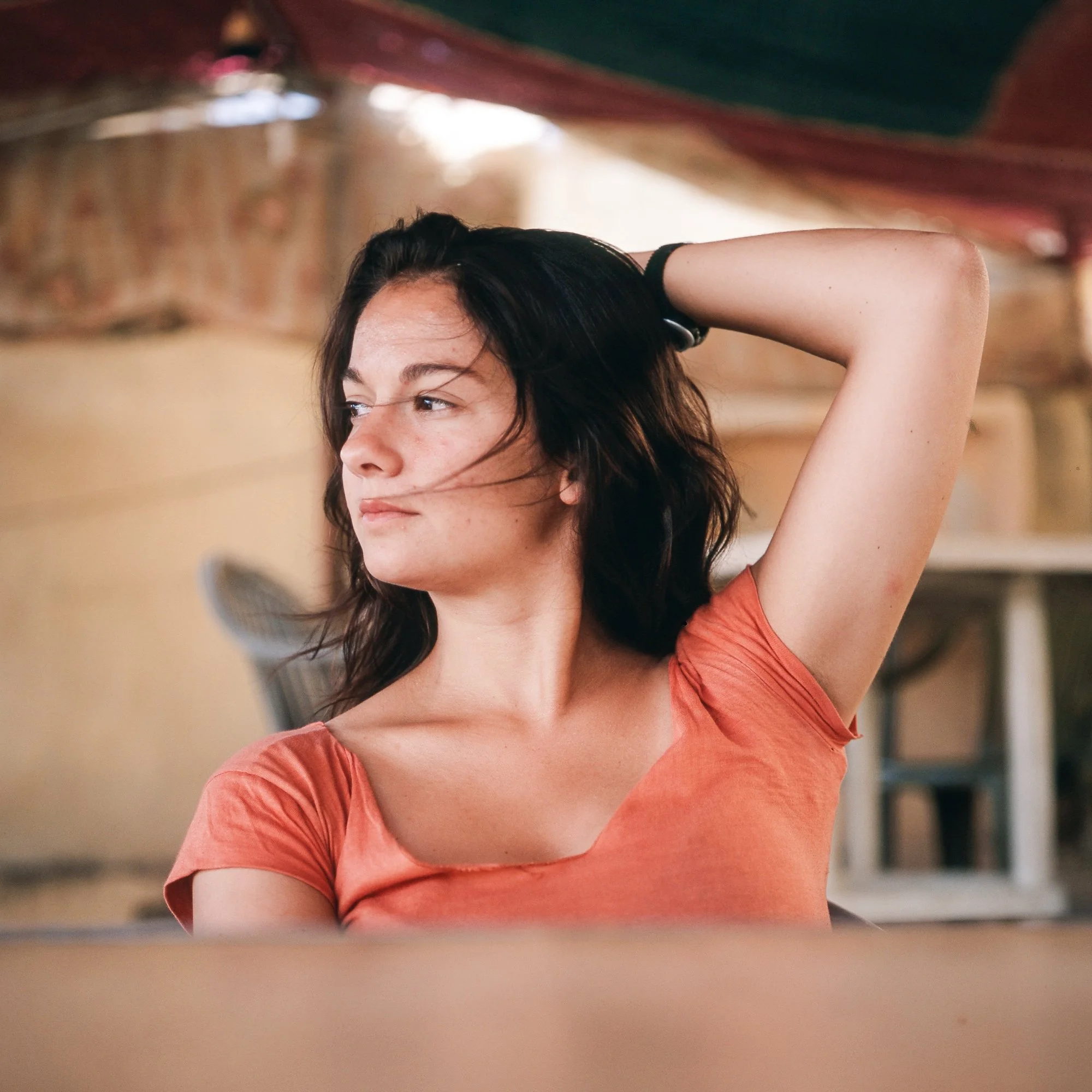 A young woman with dark hair and fair skin, wearing an orange shirt, sitting indoors with her left arm raised behind her head, looking to the left.