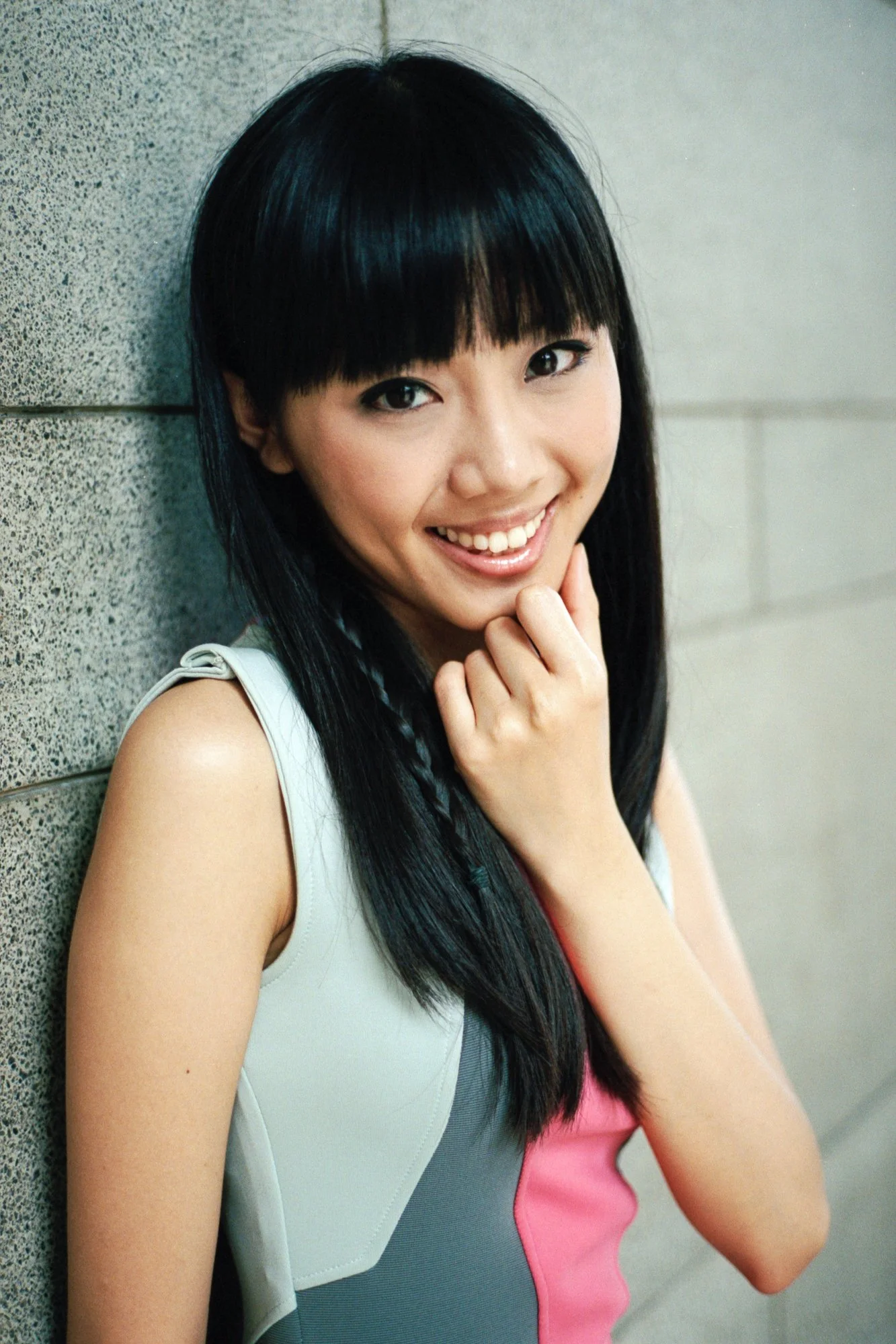 Young woman with straight black hair, smiling, leaning against a gray wall, wearing a sleeveless top with pink and gray colors.