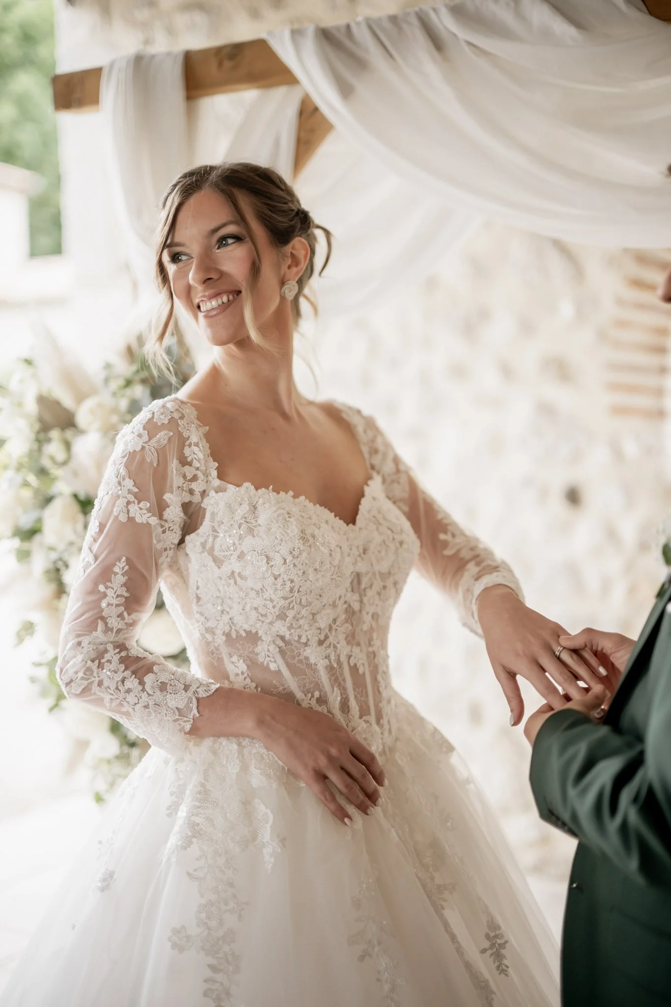 Une mariée en robe de mariage en dentelle échange un anneau avec son partenaire lors d'une cérémonie nuptiale, dans un décor lumineux avec des drapes blancs et des fleurs.
