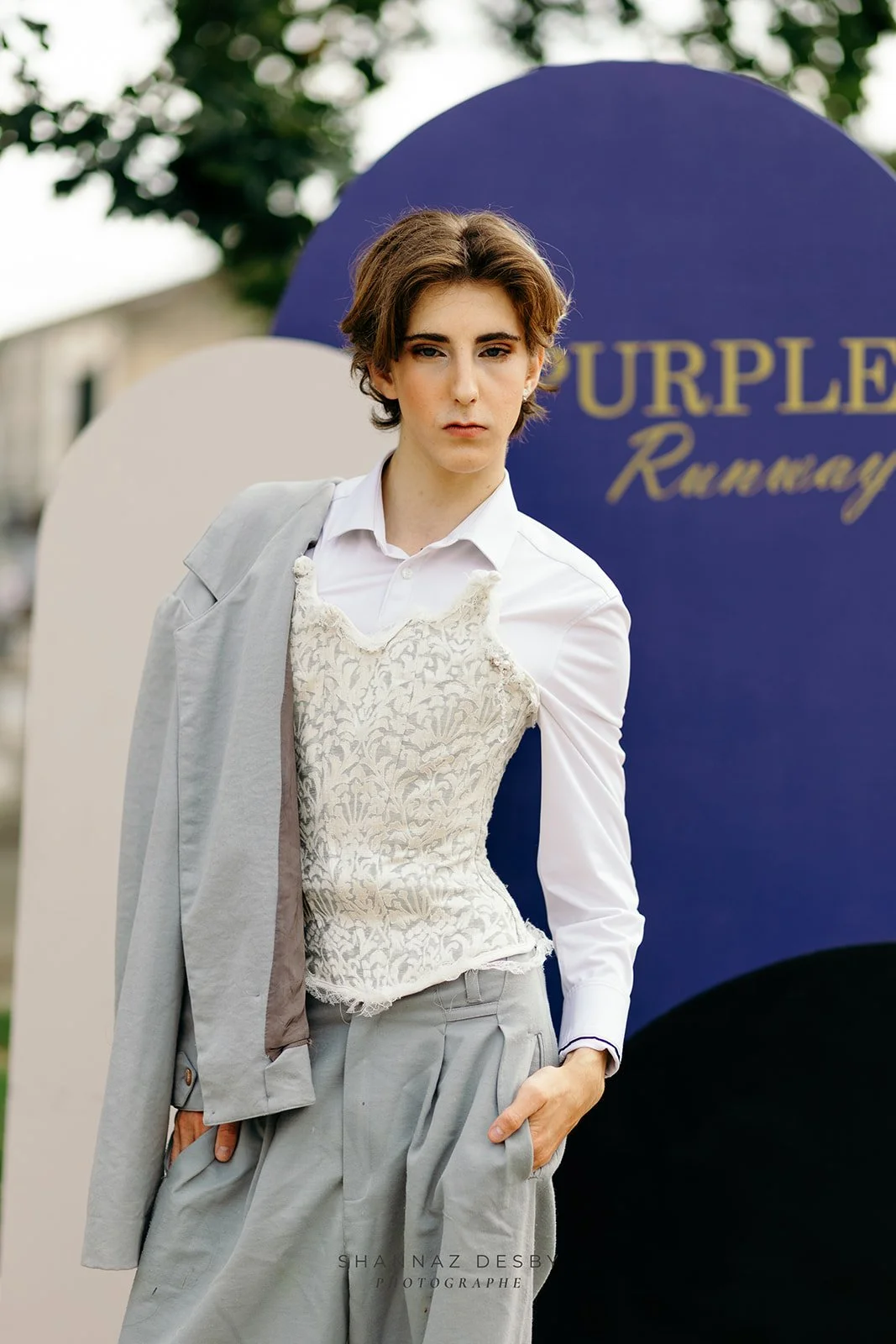 Une jeune femme avec des cheveux courts et bouclés, portant une chemise blanche, un gilet en dentelle beige, et un pantalon gris. Elle se tient devant des panneaux de couleur violet et beige avec le mot "Purple" visible sur le panneau violet.