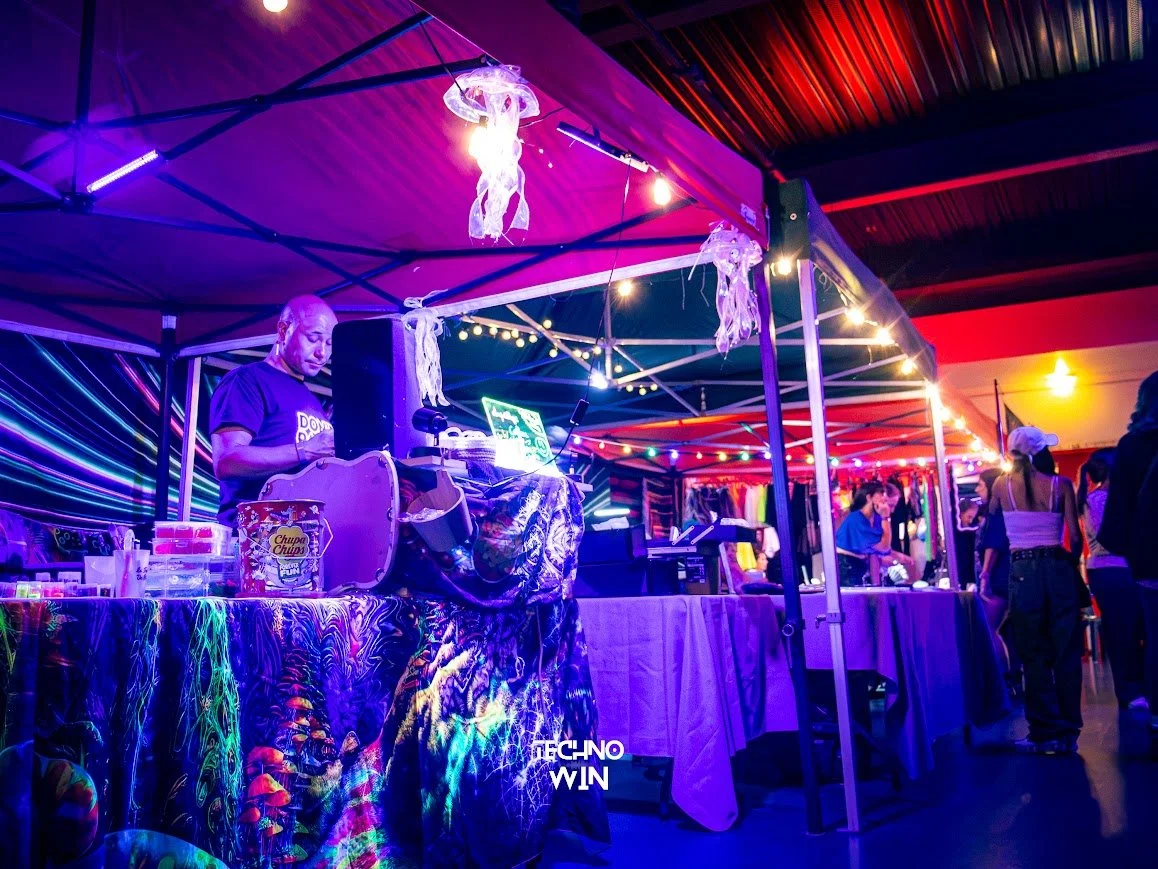 Festival ou fête en extérieur avec un DJ sous un stand de fête illuminé de lumières colorées et décorations de jellyfish en plastique suspendues. Il y a des personnes en arrière-plan et des tables recouvertes de tissus colorés.
