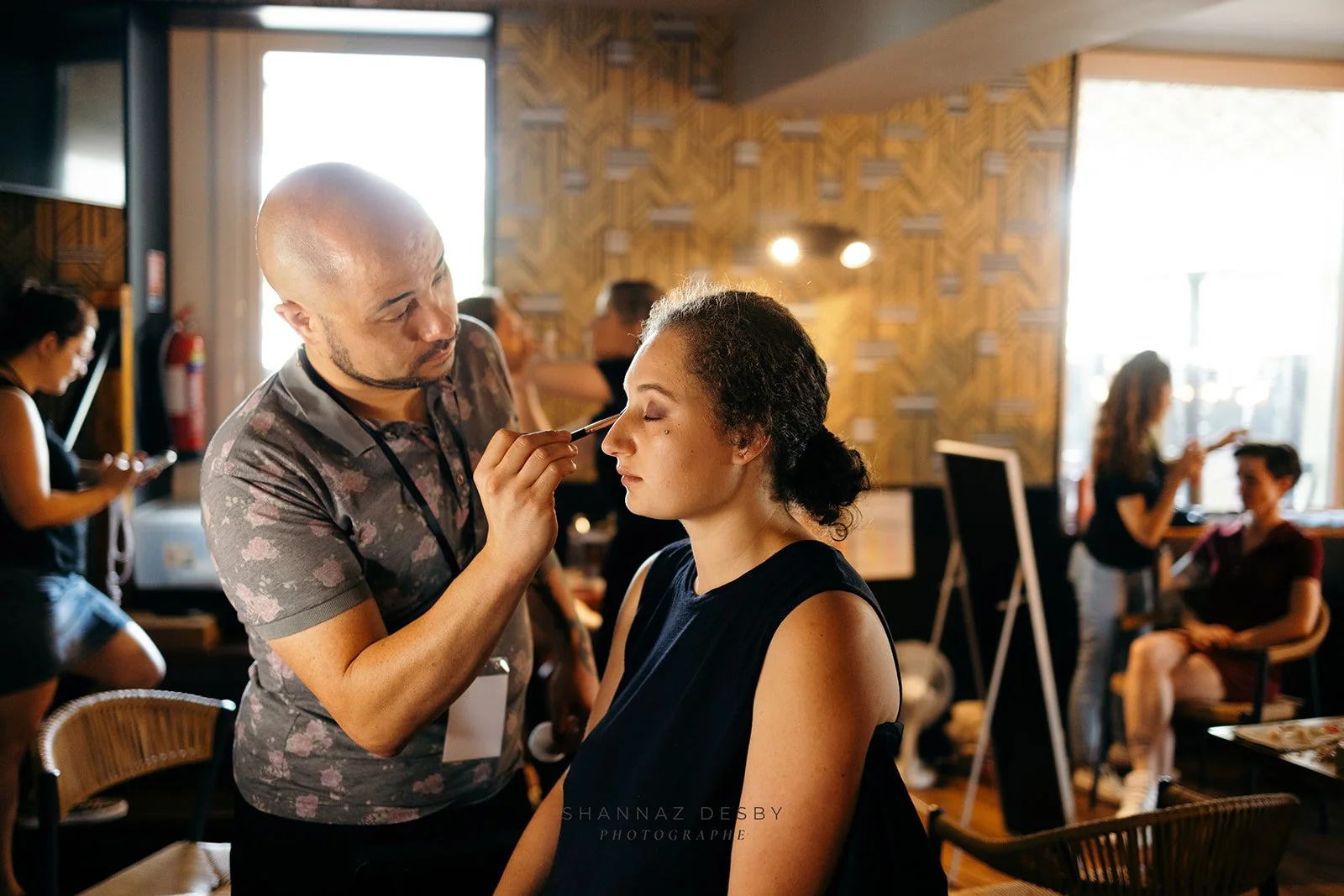 Une femme se fait maquiller par un artiste maquilleur dans un café ou un salon de beauté, avec plusieurs autres personnes en arrière-plan, assises ou utilisant leur téléphone.