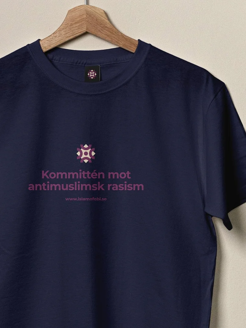 Mörkblå t-shirt med texten 'Kommunitén mot antislamisk rasism' och webbadressen 'www.islamofobi.se' i rosa, hängd på en trägalge.