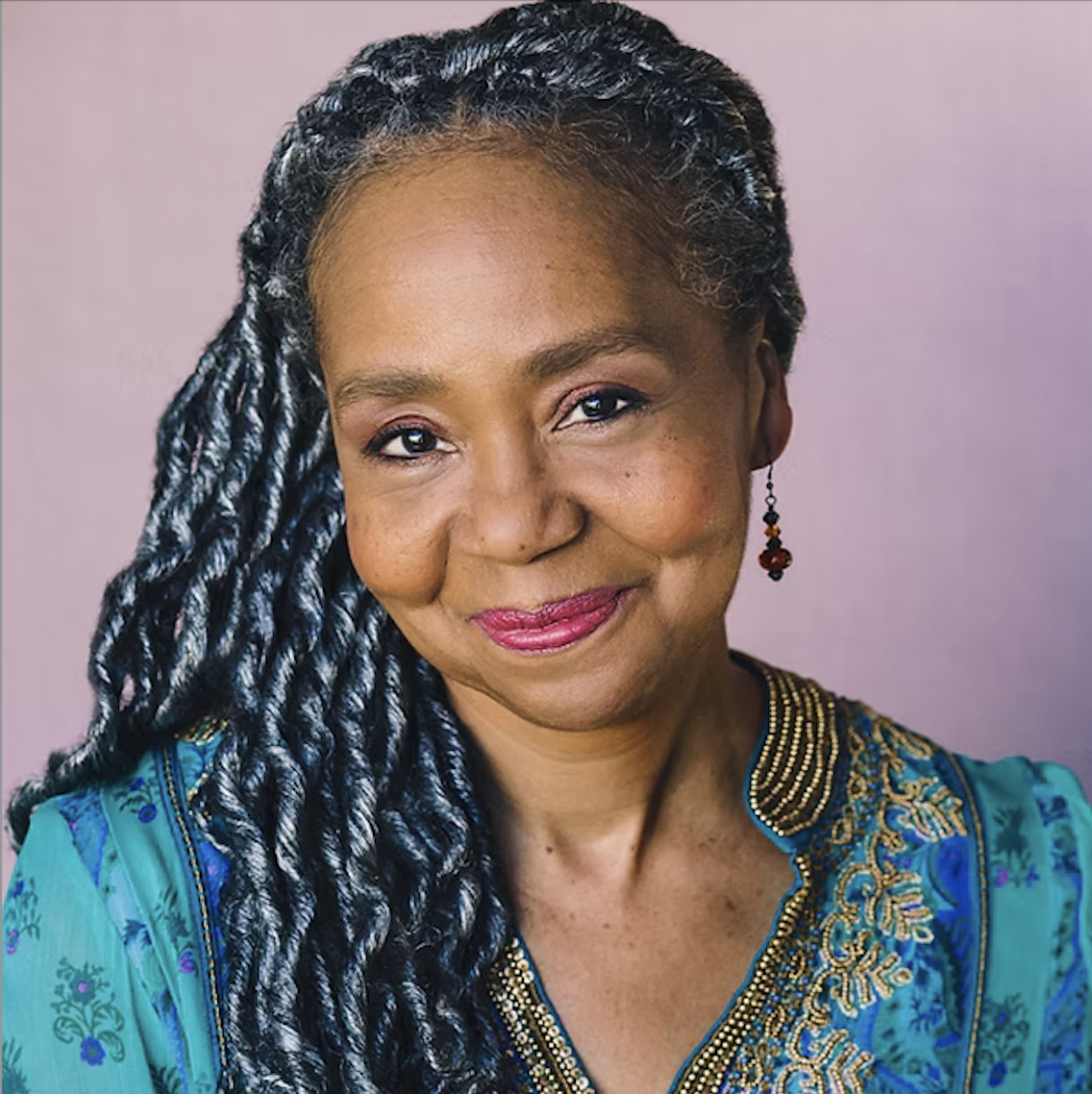 A woman with medium brown skin and braided hair, wearing a blue traditional dress with gold embroidery and red earrings, smiling against a pink background.