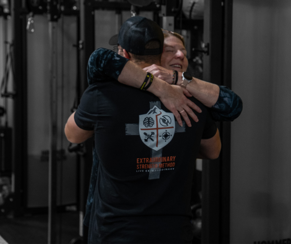 Two people hugging, one facing away wearing a black cap and T-shirt, the other smiling with eyes closed, in a gym setting.