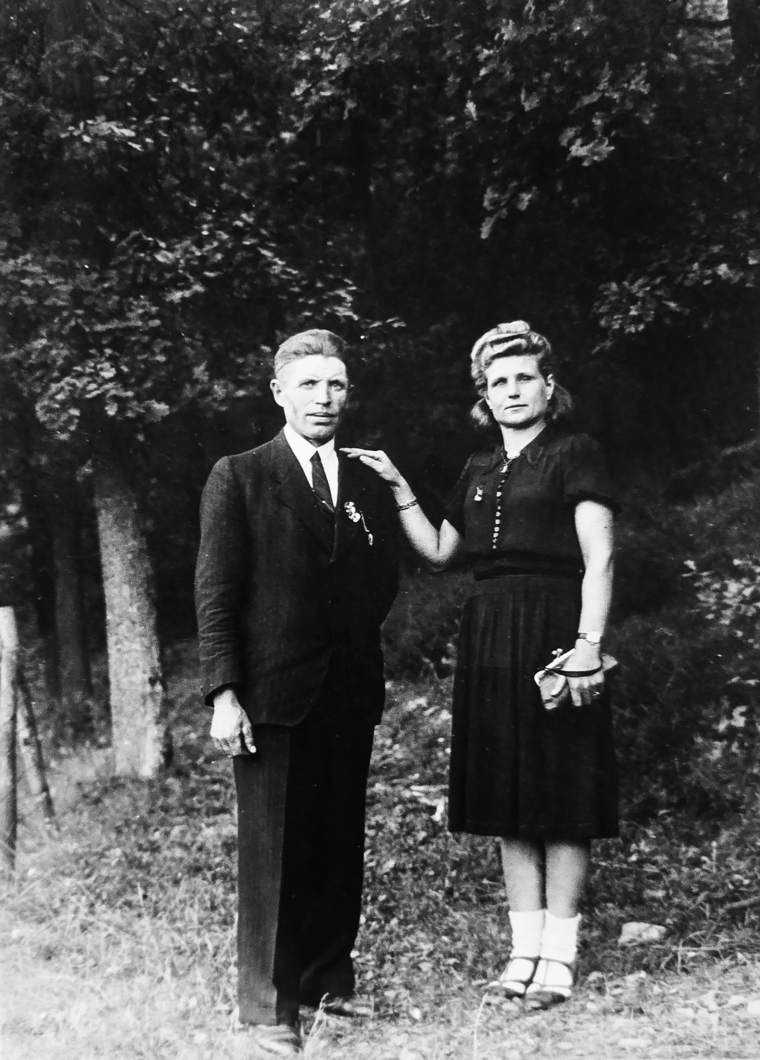 Black and white portrait of a man in a suit and a woman in a dress standing outdoors in front of trees.
