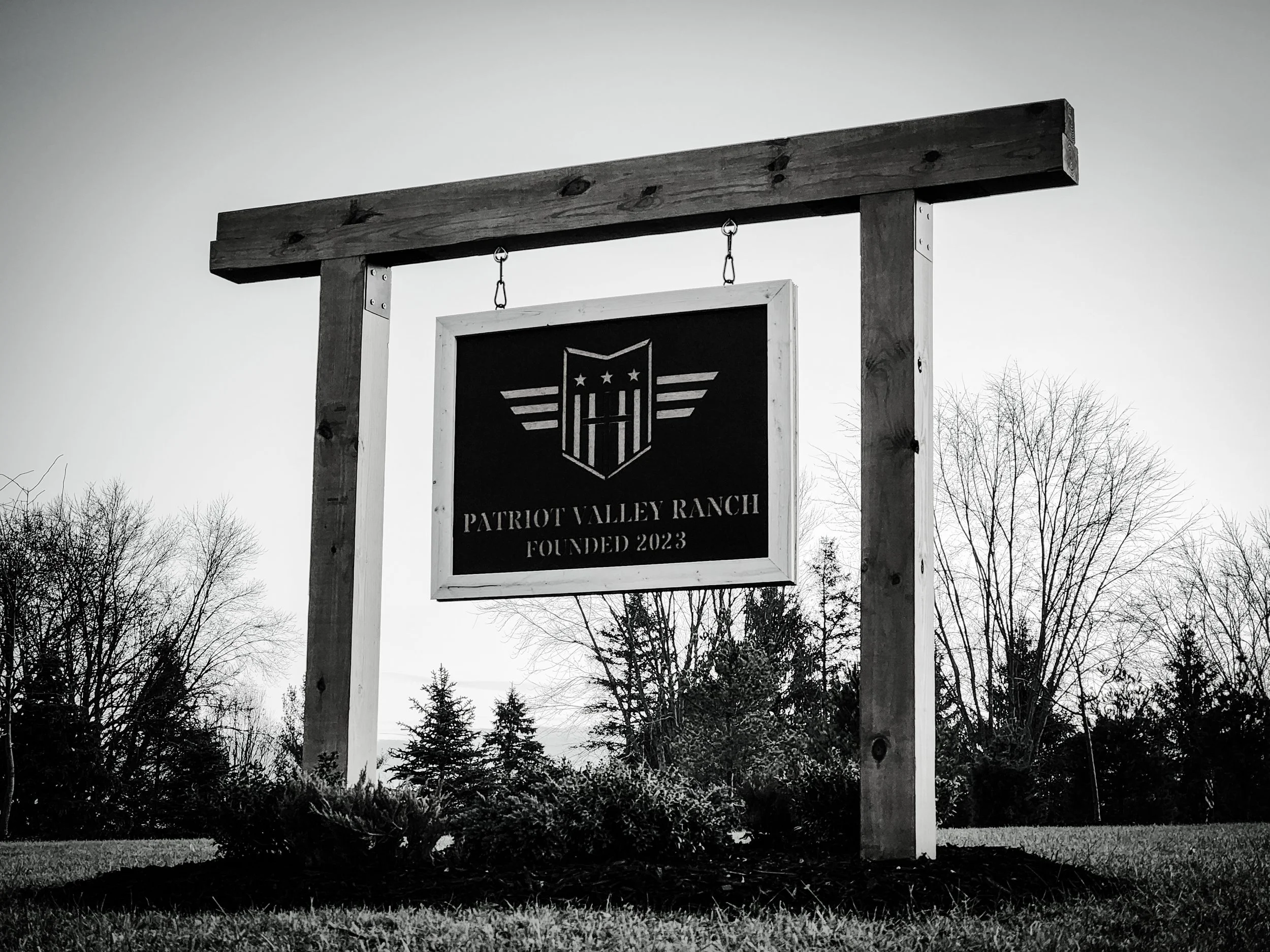 Sign reading 'Patriot Valley Ranch Founded 2023' hanging from wooden frame, outdoors with trees and grass.