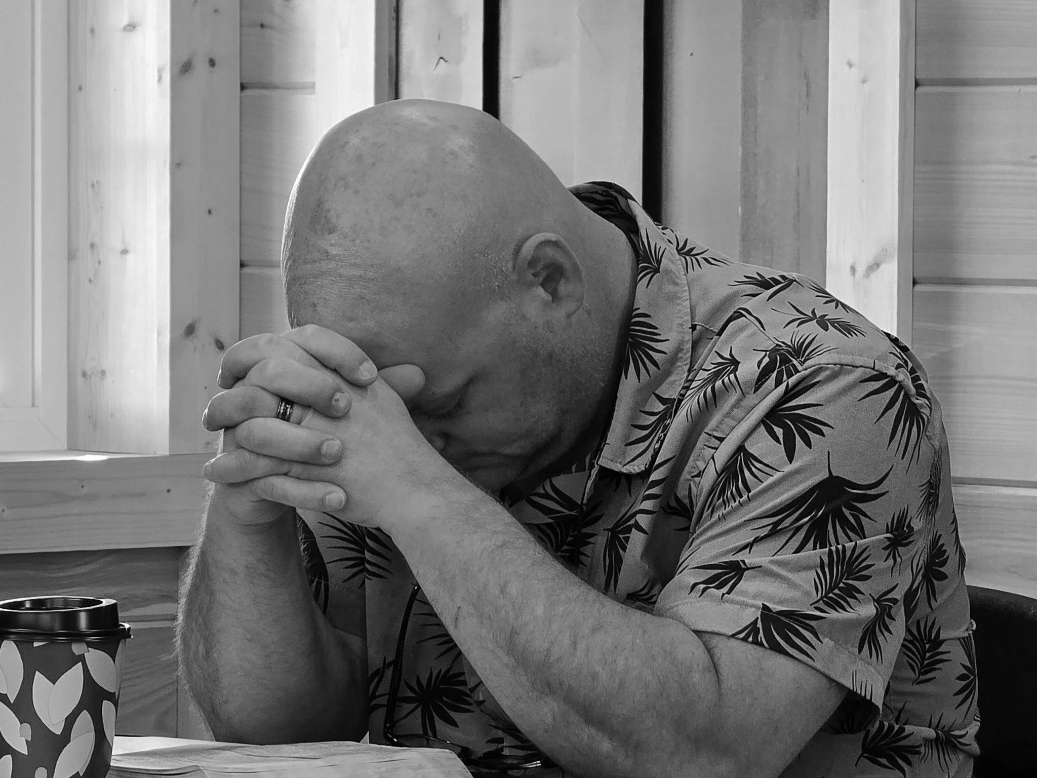 A bald man resting his head on his arms with closed eyes, sitting at a wooden table in a room with light wood-paneled walls. There is a coffee cup and a reusable travel mug on the table.