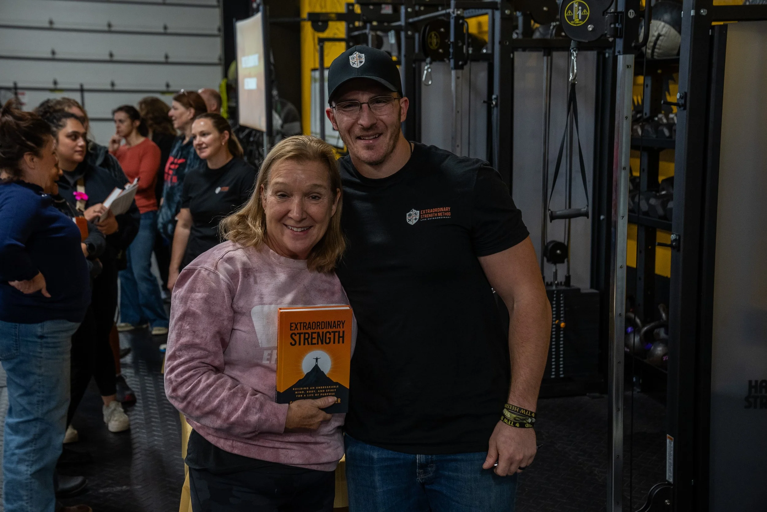 A woman holding a book titled 'Extraordinary Strength' standing next to a man, both smiling, in a gym with people in the background.