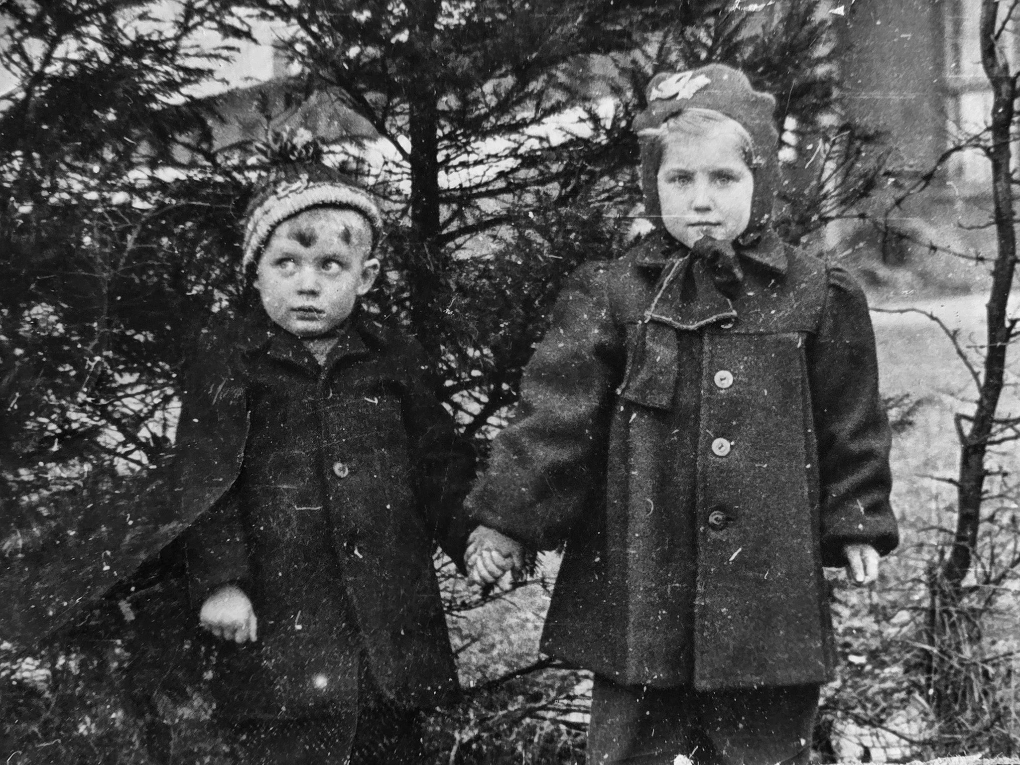 Two children, a boy and a girl, holding hands outdoors near trees, wearing dark coats, in black and white.