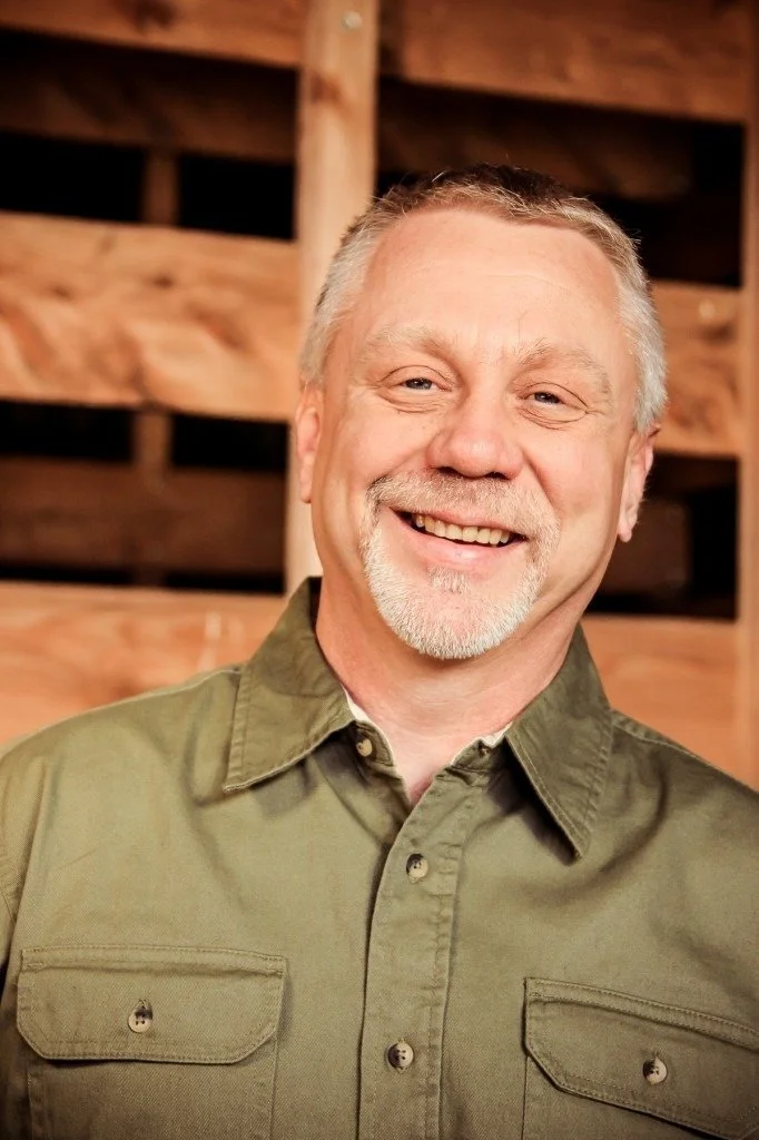 A smiling middle-aged man with short gray hair, beard, and mustache wearing an olive-green button-up shirt, standing in front of a wooden background.
