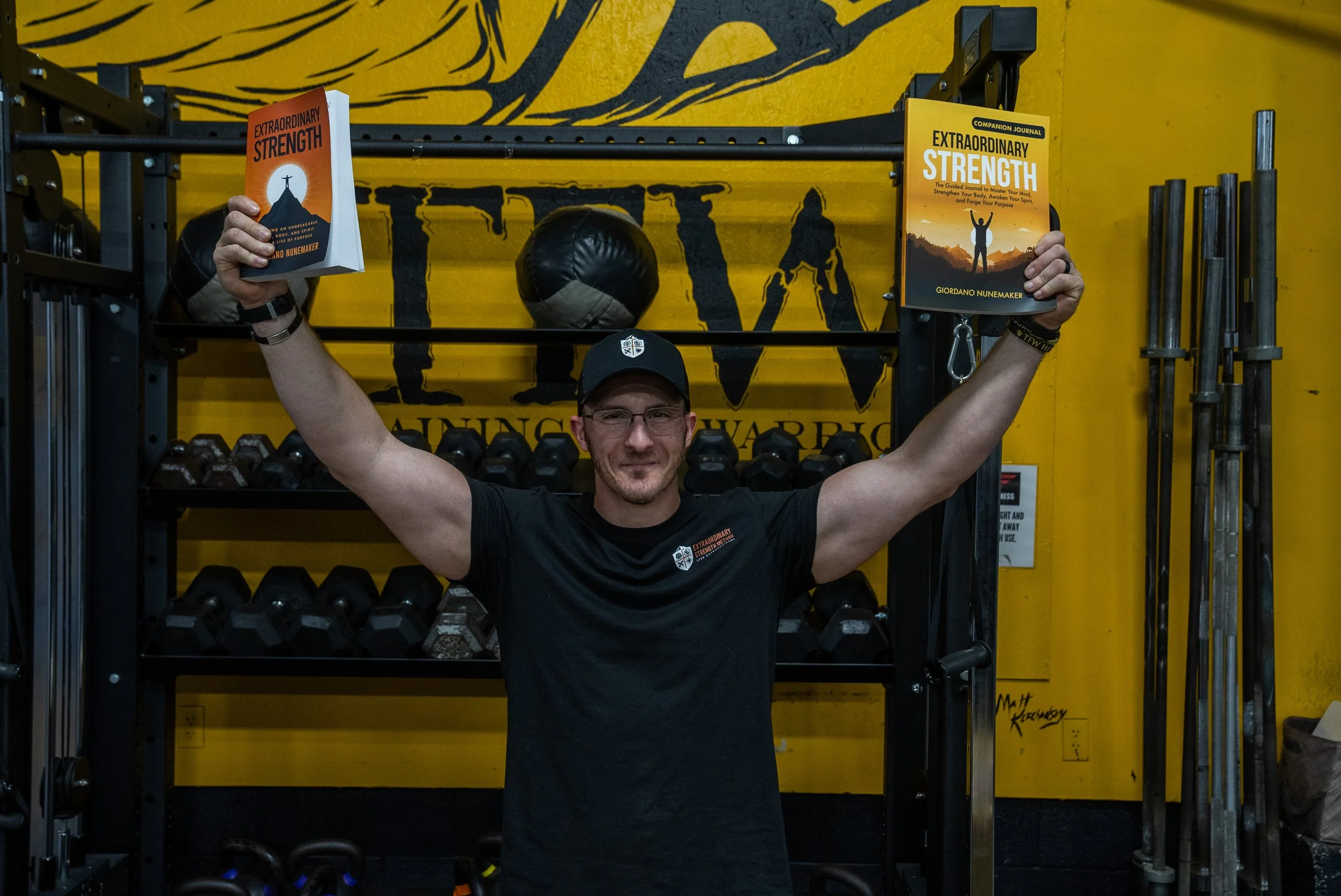 A man wearing glasses, a black cap, and a black T-shirt, standing in a gym with yellow walls, holding two copies of a book titled 'Extraordinary Strength' by Giordano NuneMaker, one in each hand raised above his shoulders.