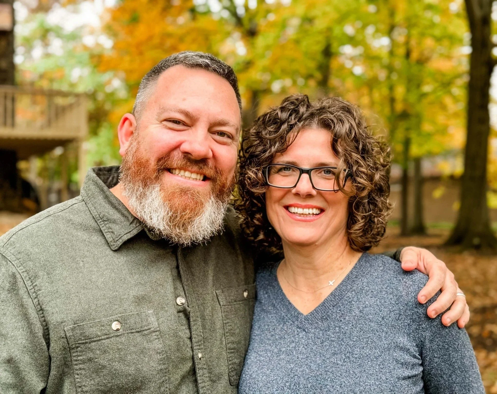 Smiling man with a beard and woman with curly hair and glasses standing outdoors in front of autumn trees.