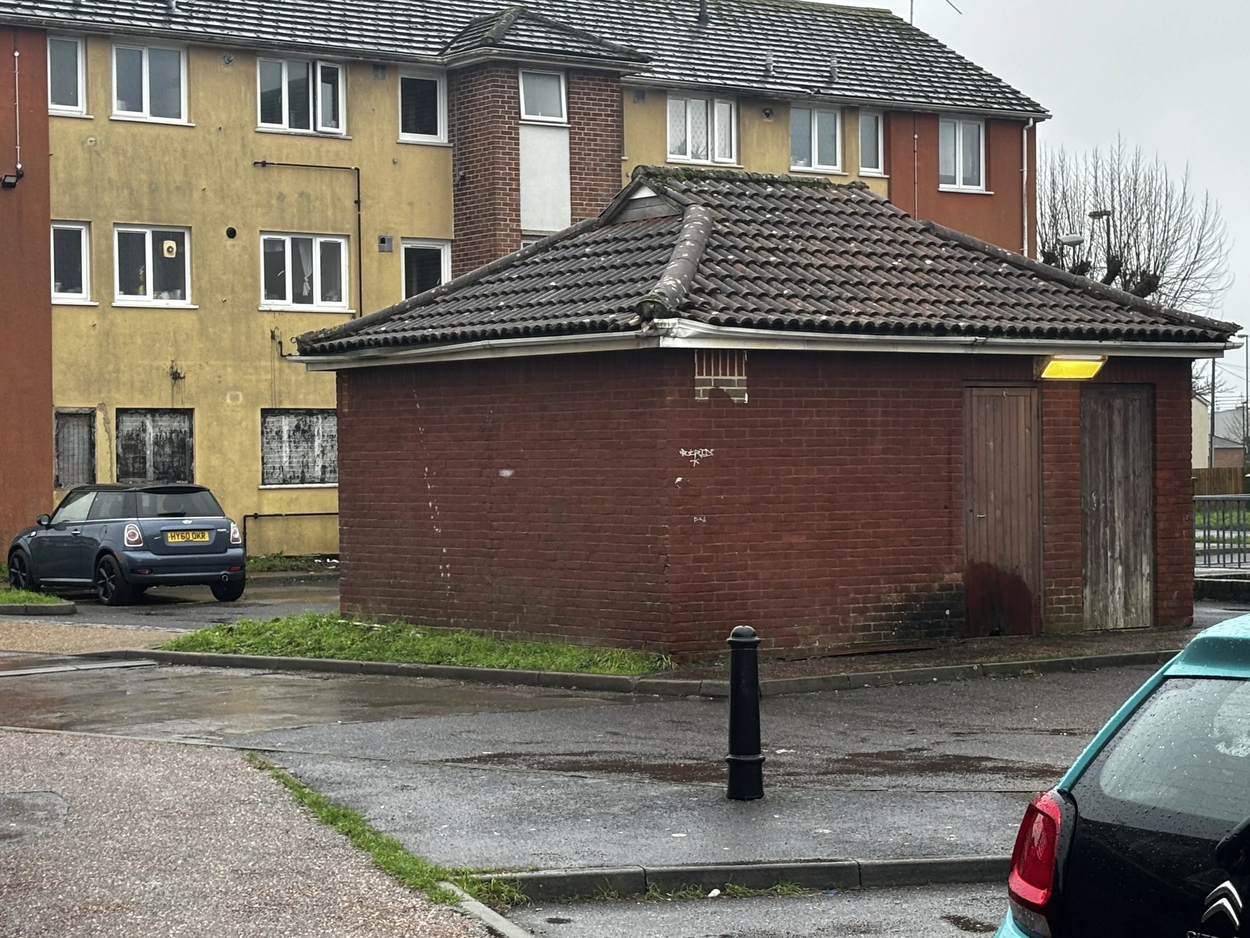 Deep brown small electricity building one level set in front of a 3 storey dark yellow block of flats all in the rain