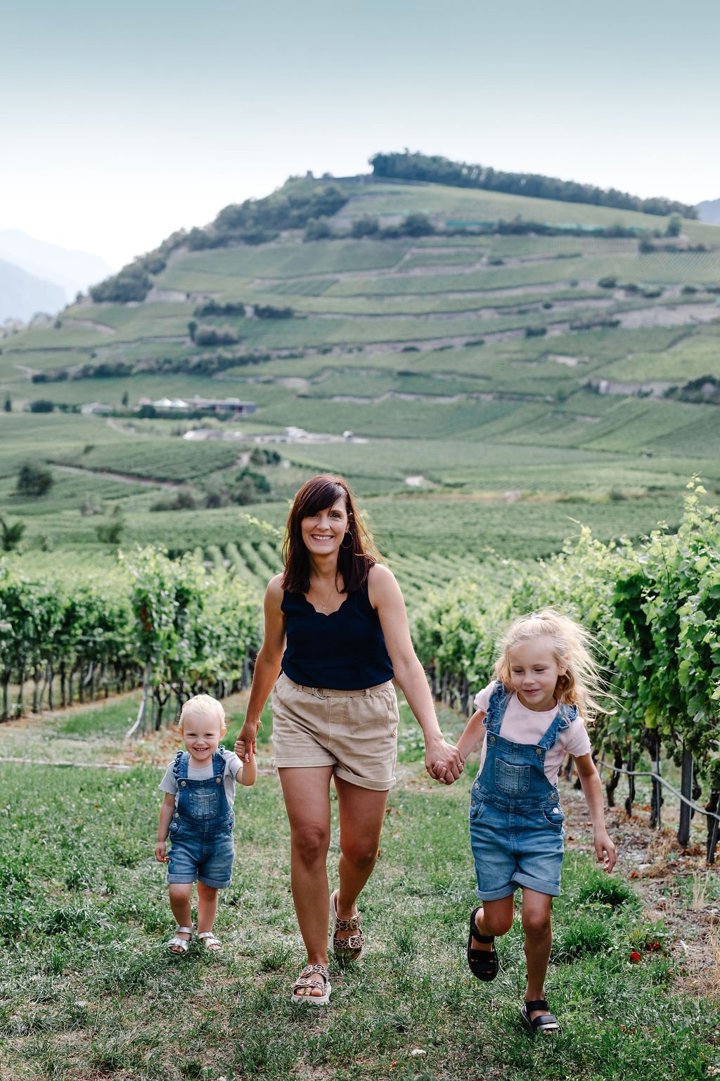 Une femme souriante avec deux jeunes enfants, une fille et un garçon, marchant dans un vignoble avec des collines verdoyantes en arrière-plan.