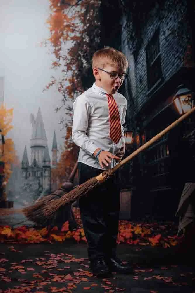 Un jeune garçon habillé en uniforme scolaire avec une cravate rayée, tenant un balai en bois, dans un décor d'automne avec des feuilles tombées, un bâtiment ancien en arrière-plan, probablement à thème Harry Potter.