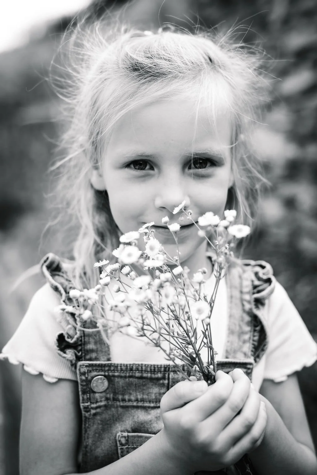 Une jeune fille blonde tient un bouquet de petites fleurs blanches devant son visage, dans une photo en noir et blanc.