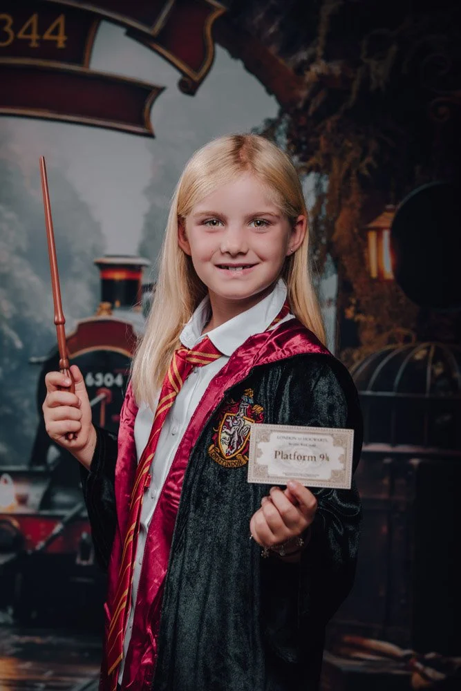 Une jeune fille habillée en sorcière de Harry Potter avec une écharpe rayée rouge et or, tenant un billet de train pour Poudlard et une baguette magique, devant une arrière-plan de train et d'une forêt enchantée.