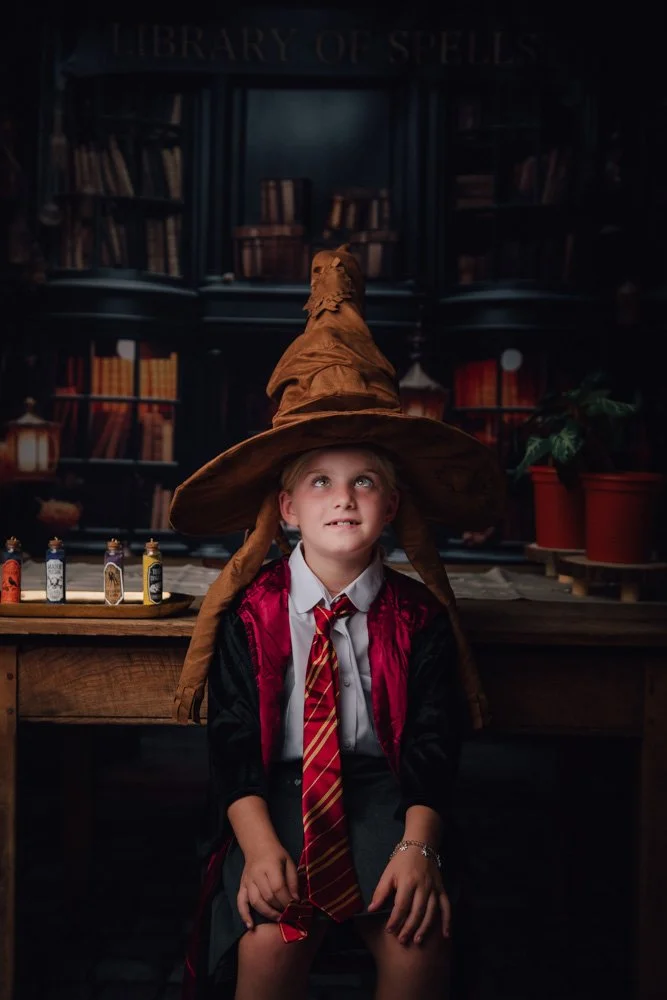 Une jeune fille portant une robe de sorcière et un chapeau pointu de sorcier assise devant une bibliothèque magique.