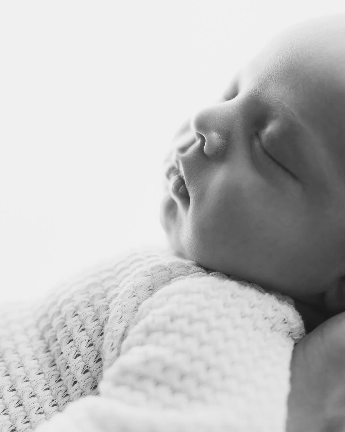 Un bébé endormi, en noir et blanc, où seul le visage est visible, posé sur une couverture en tricot. Photo faite au studio de photographie pour nouveau-né au Viens et Vois en Valais