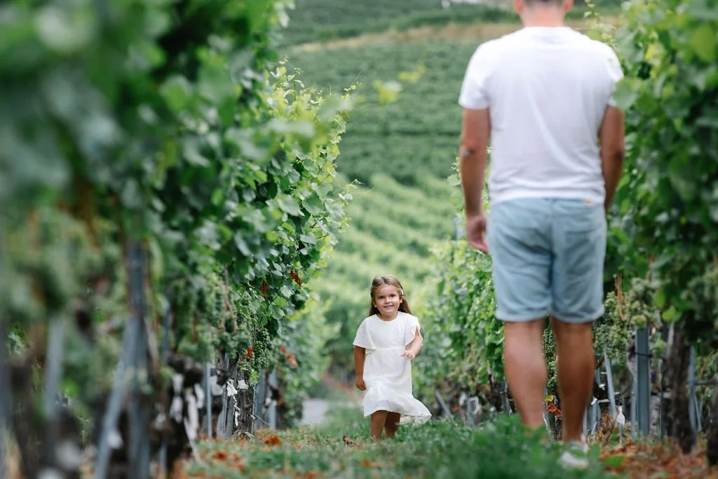 Une petite fille souriante portant une robe blanche marche dans un vignoble pendant qu'un homme, probablement son père, marche devant elle.