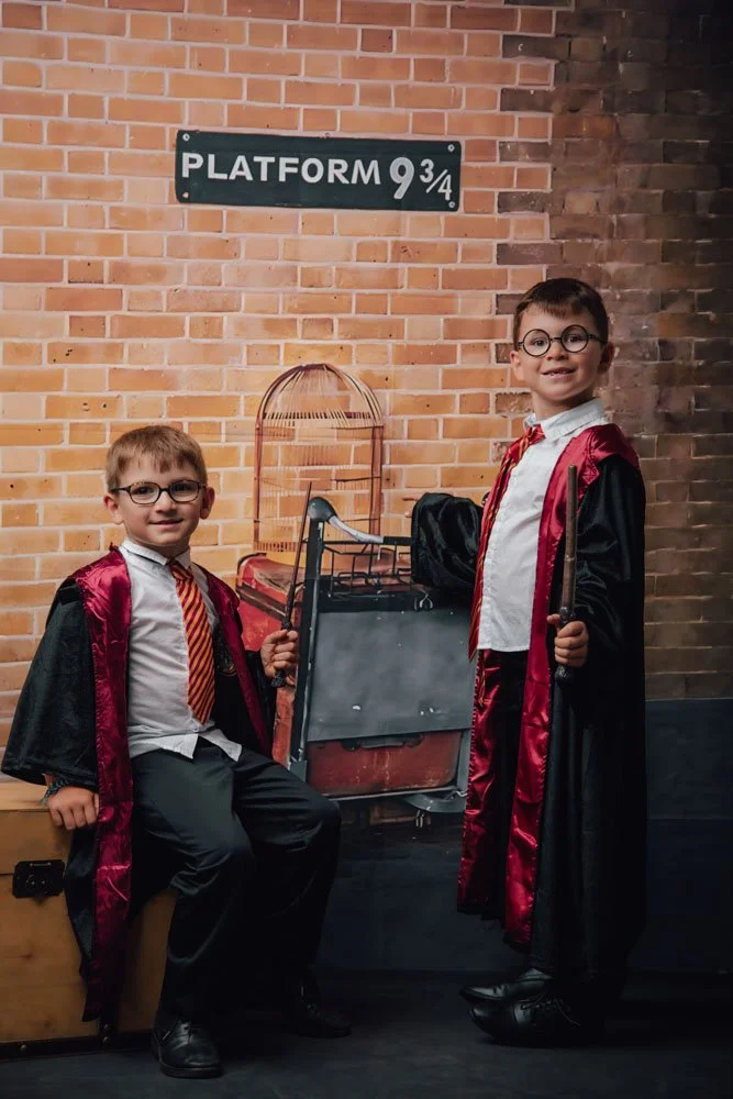 Deux enfants habillés en sorciers avec des robes noires et des costumes cravate rouges, posant avec une valise en forme de chariot de train à côté d'une affiche 'Plateforme 9¾' sur un mur en briques.