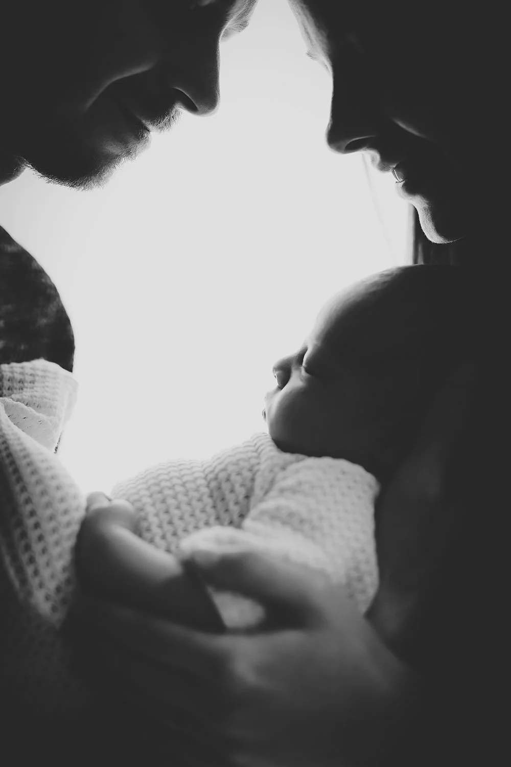 Une famille regarde tendrement un bébé dans une photo en noir et blanc. Photo faite au studio de photographie pour nouveau-né au Viens et Vois en Valais