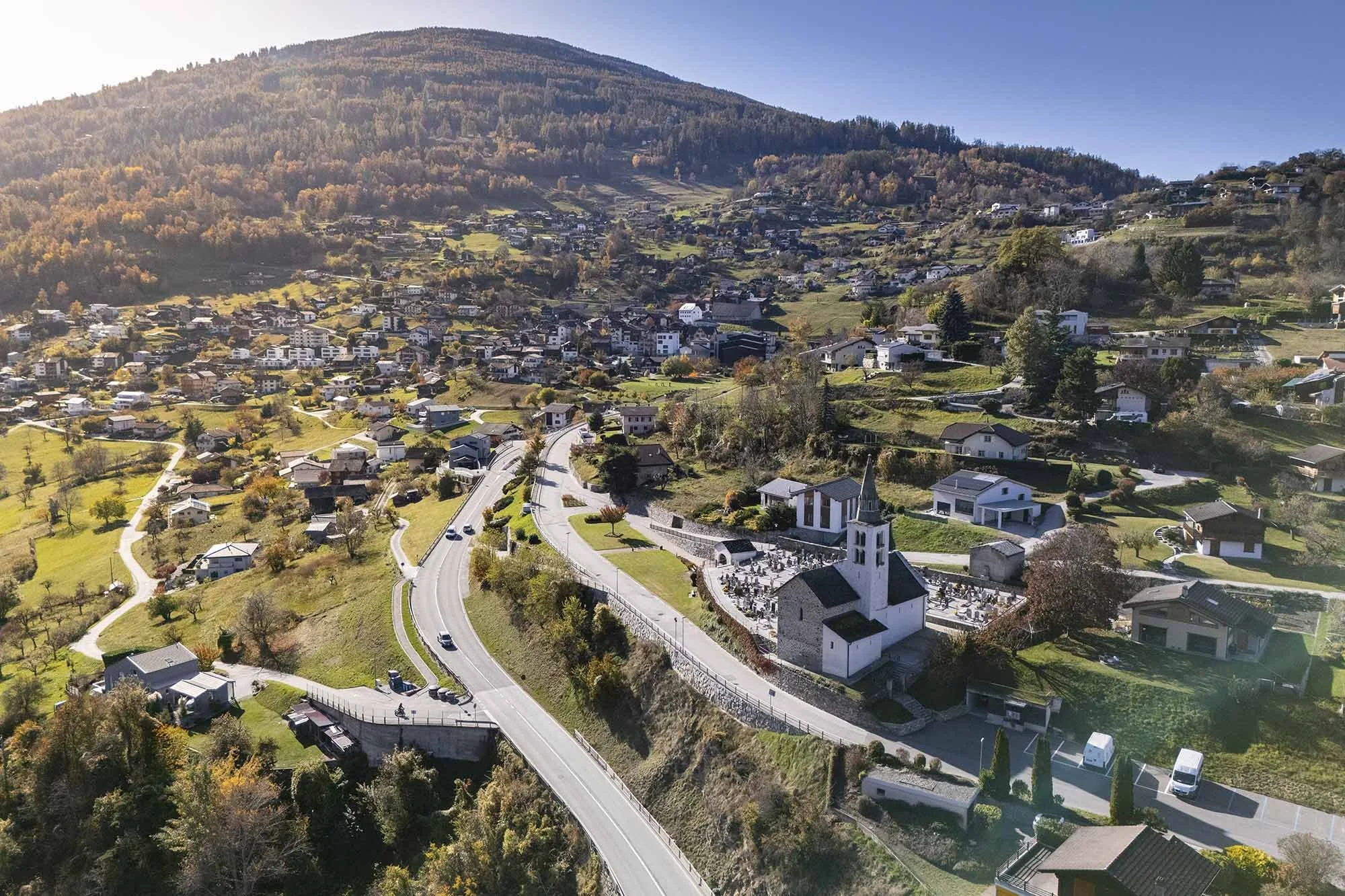 Vues aériennes d'un village de montagne avec une église blanche au centre, entourée de maisons et de routes sinueuses, avec une forêt et une montagne en arrière-plan en automne.