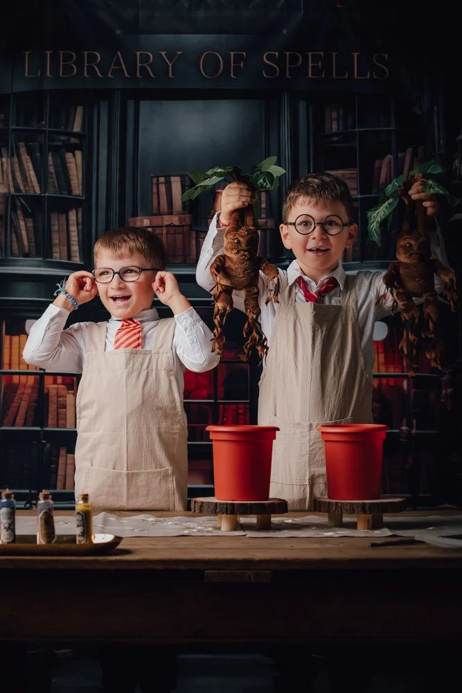 Deux jeunes garçons déguisés en sorciers avec des lunettes, portant des tabliers, dans une bibliothèque magique, jouent avec des racines de poupée ou des figurines de sorciers, avec des pots en terre cuite et des fioles sur la table.