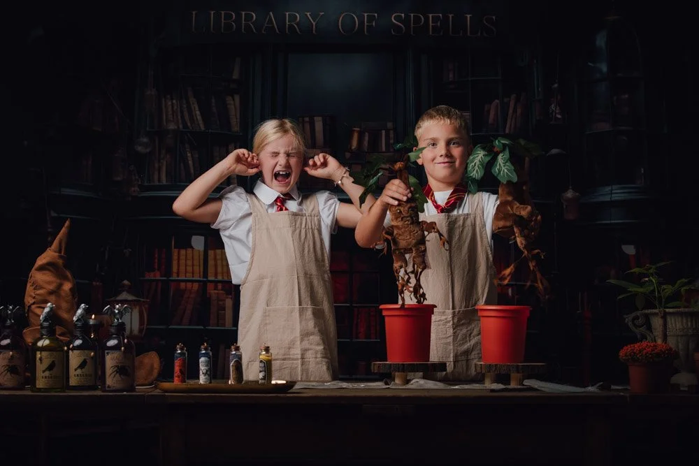 Deux enfants en blouse d'apprenti sorcier dans une bibliothèque de sorts, l'un souriant et l'autre criant, tenant des plantes dans des pots de fleurs.