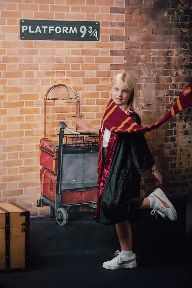 Une jeune fille déguisée en personnage de Harry Potter, portant une robe de sorcier, un foulard aux couleurs de Gryffondor, et des baskets blanches, posant avec une expression amusante devant un mur en briques et un panneau indiquant la plateforme 9