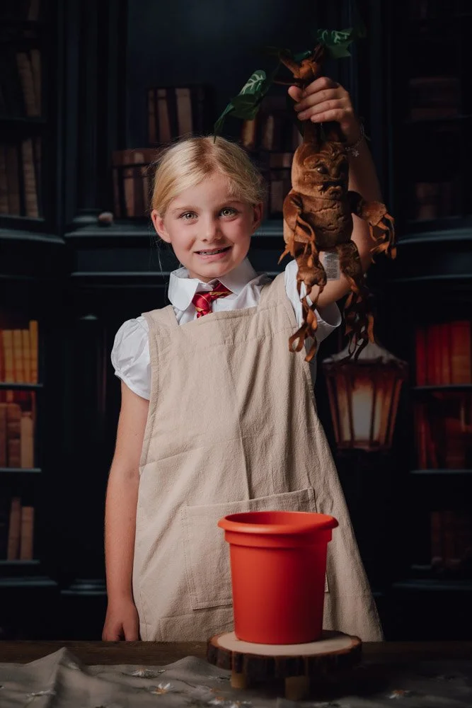 Une jeune fille souriante, portant un tablier beige et une cravate rouge, tient une grenouille en hauteur dans un laboratoire ou une salle de classe, avec un pot en terre cuite rouge sur une table en bois devant elle.