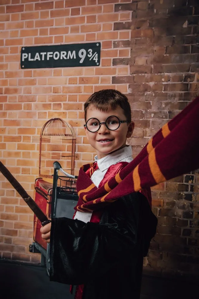 Jeune garçon déguisé en Harry Potter avec un écharpe Gryffondor, lunettes rondes, robe de sorcier, tenant une baguette, devant un mur en briques avec une enseigne 'Plateforme 9 3/4'.