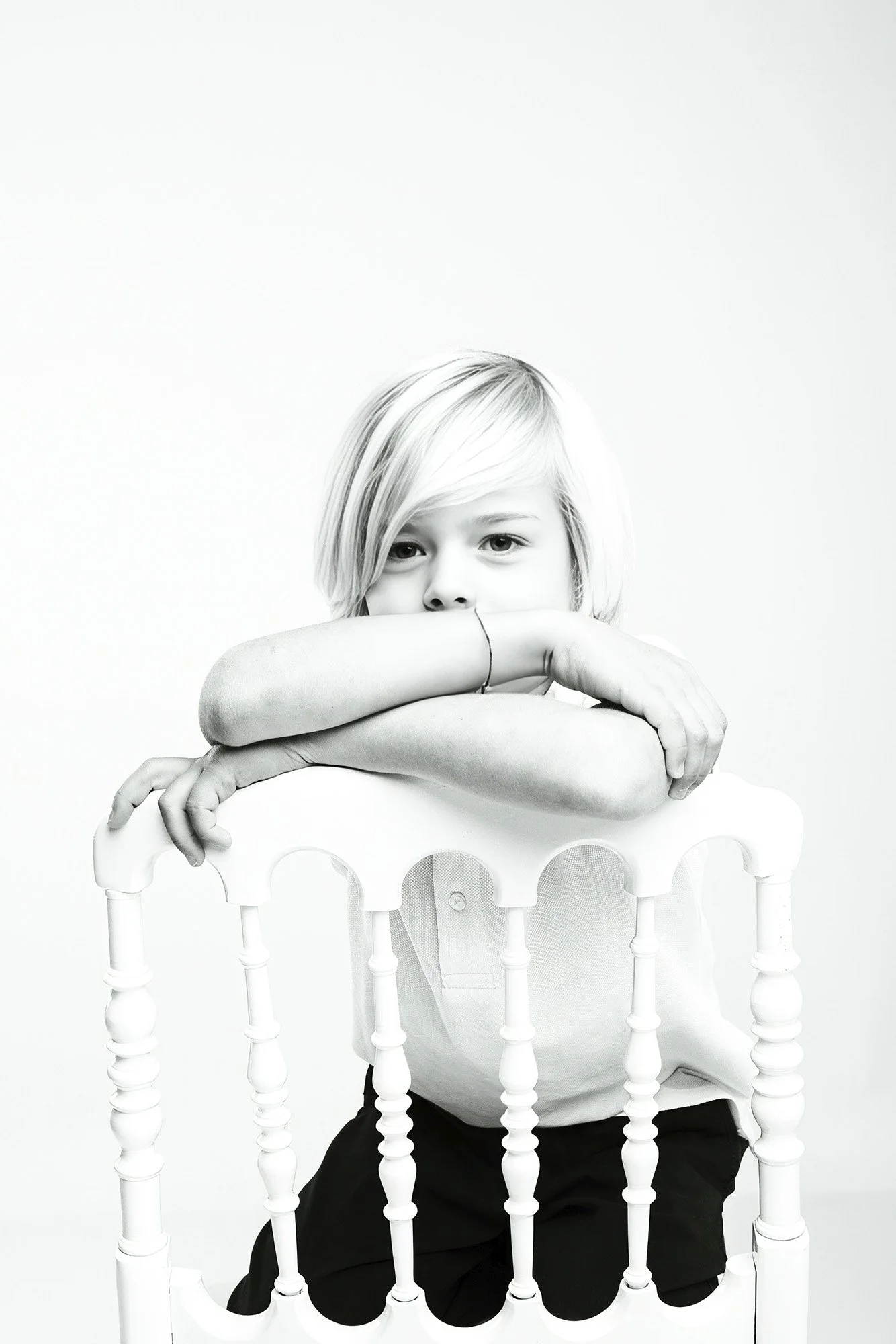 Une fille aux cheveux blonds, assise dans une chaise en plastique blanche, repose ses bras sur le dossier et regarde l'objectif. L'image est en noir et blanc, le fond est blanc.