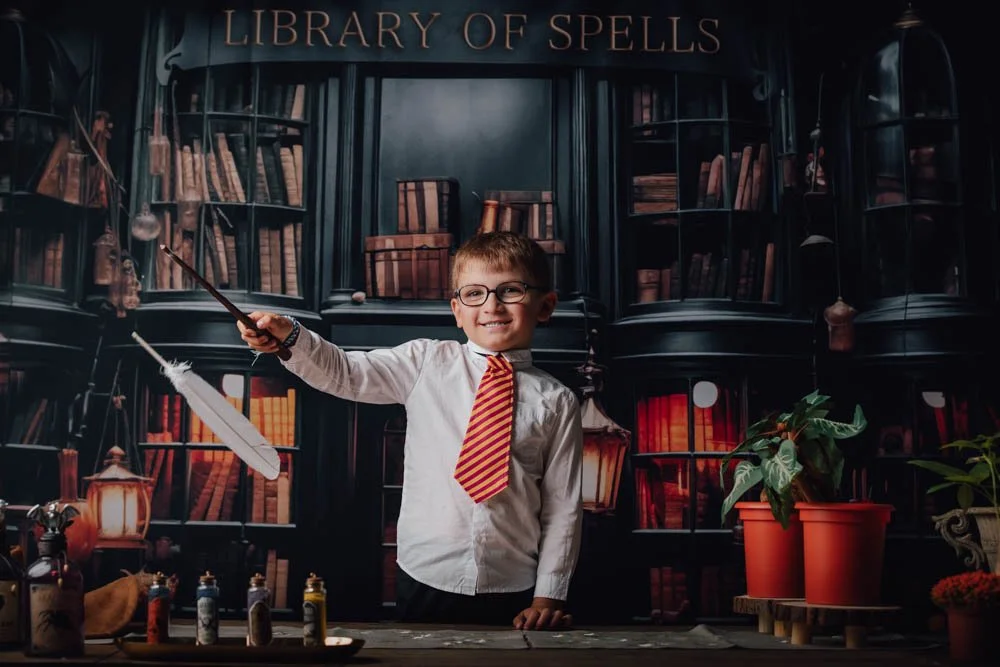 Un jeune garçon souriant, portant une chemise blanche, une cravate rayée rouge et jaune, et des lunettes, se tient devant une étagère avec le panneau "Library of Spells". Il tient une plume blanche dans une main et un bâton dans l'autre, évoquant un