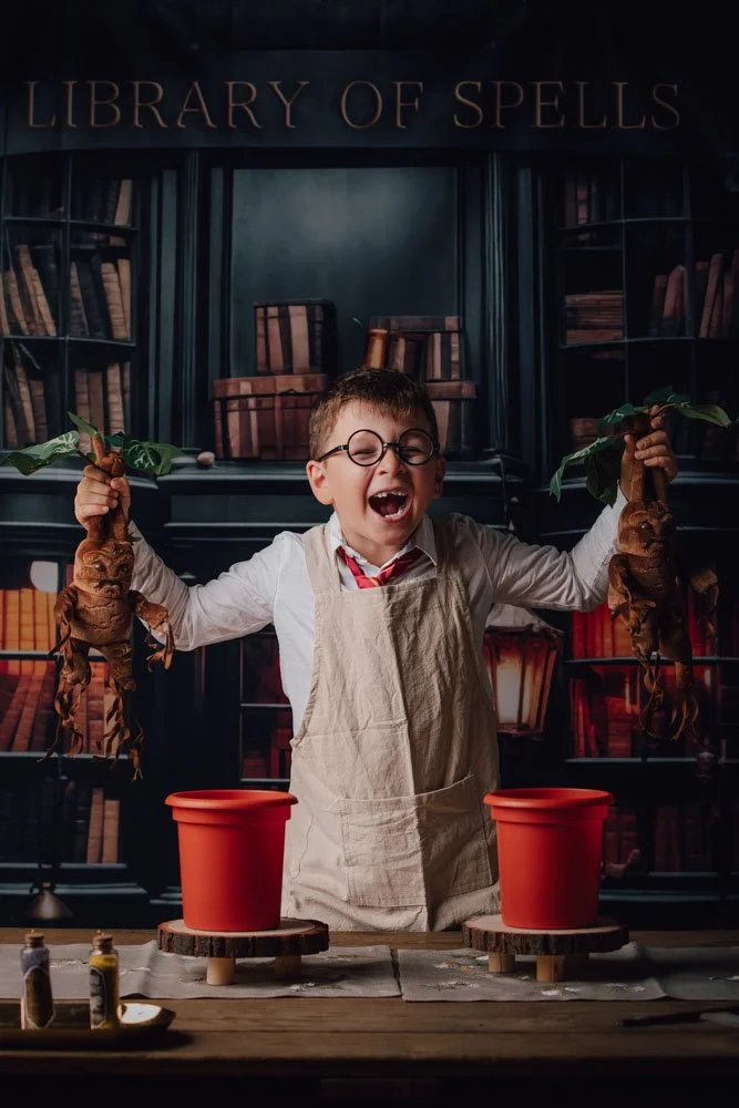 Un garçon avec des lunettes et un tablier beige, souriant et levant deux racines de mandragore dans un décor de bibliothèque magique avec une enseigne "Library of Spells".