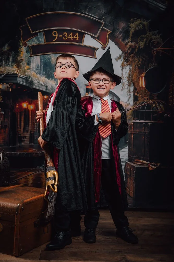 Deux enfants habillés en costumes de sorciers devant un décor de chemin de fer magique, un d'eux porte une robe noire avec une baguette, l'autre un costume avec un chapeau pointu et une baguette.